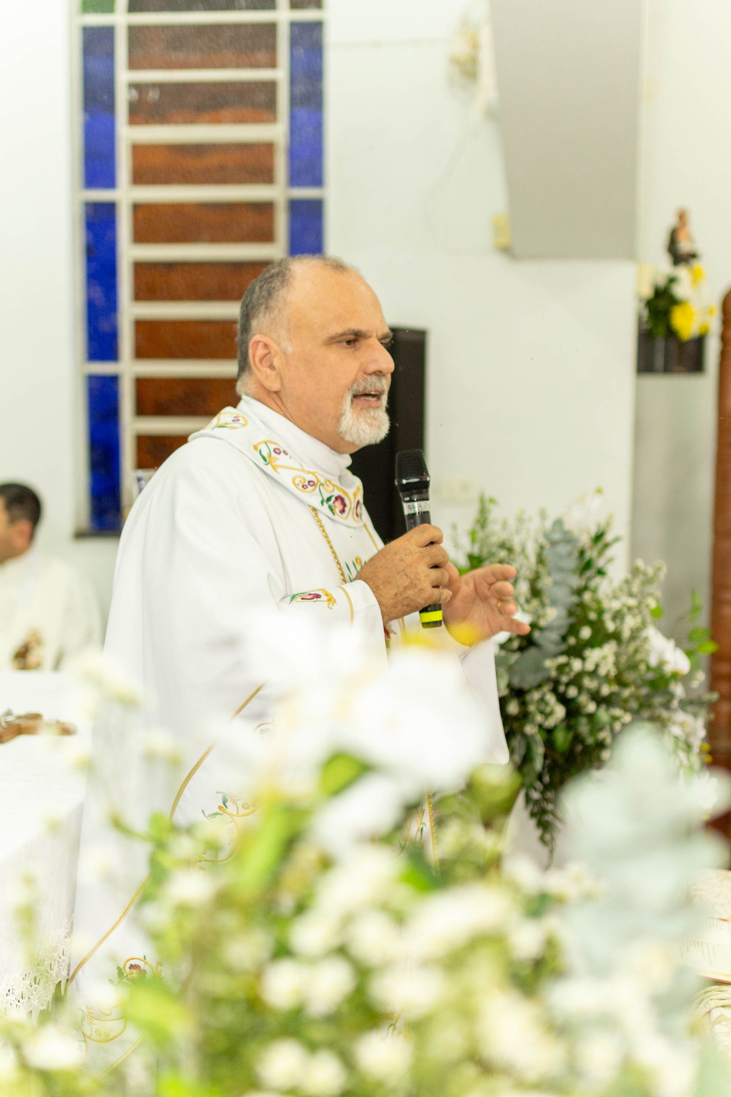 Catholic priest in white vestments speaks into microphone at altar adorned with white flowers during Mass ceremony.