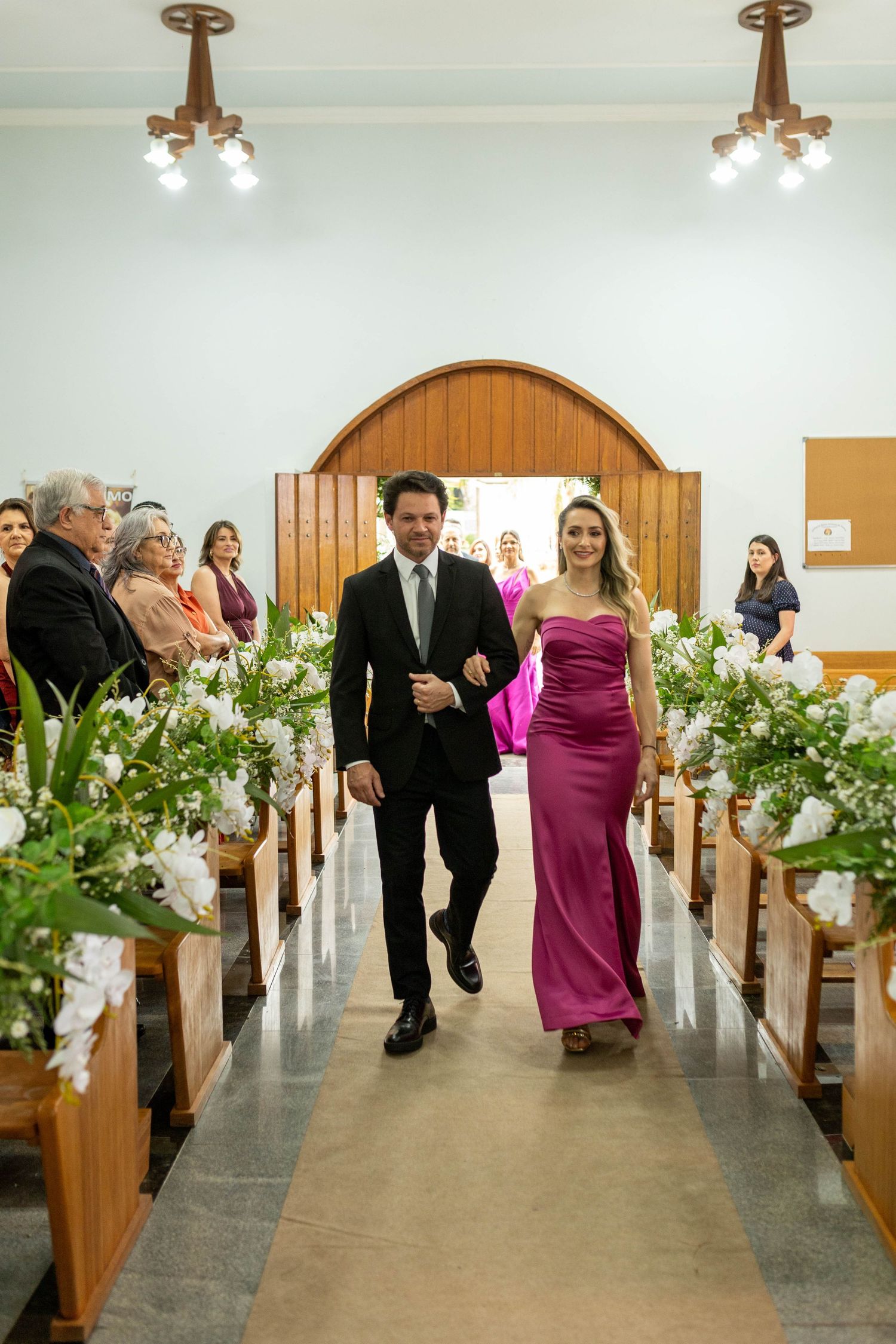 A man in a suit and woman in a pink gown walk down a flower-lined church aisle during a wedding ceremony.