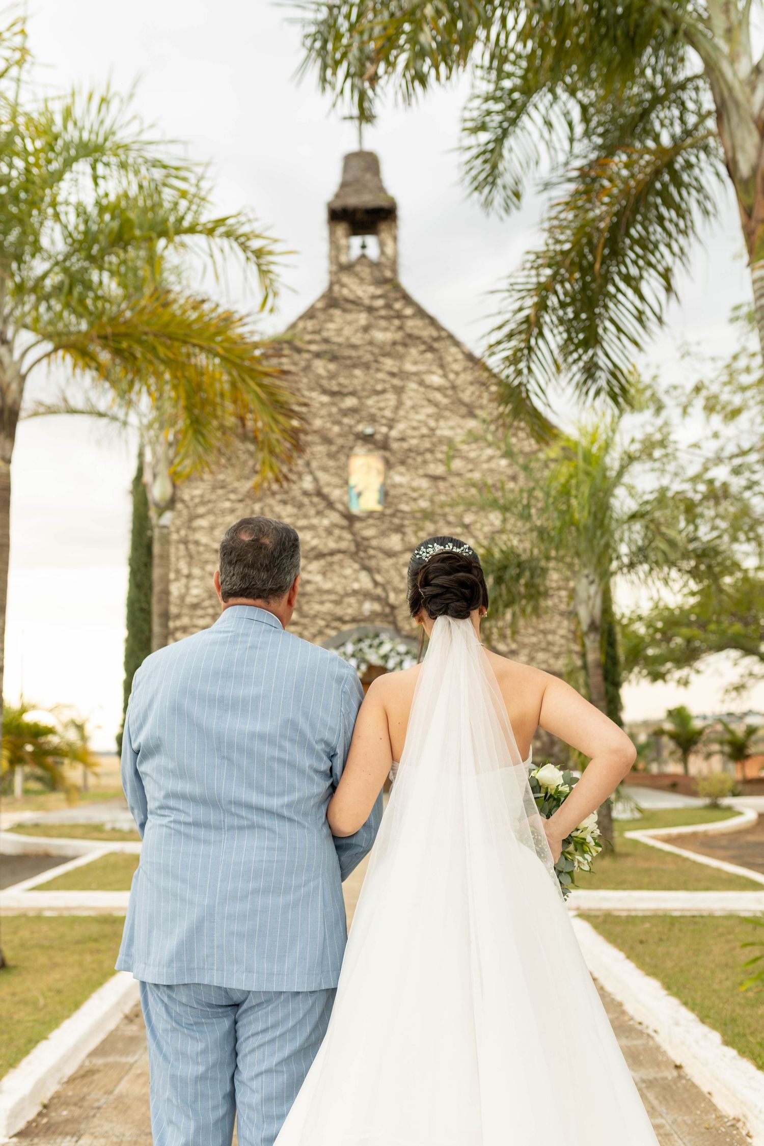Bride in white gown and veil walks with man in blue suit toward stone chapel surrounded by palm trees.