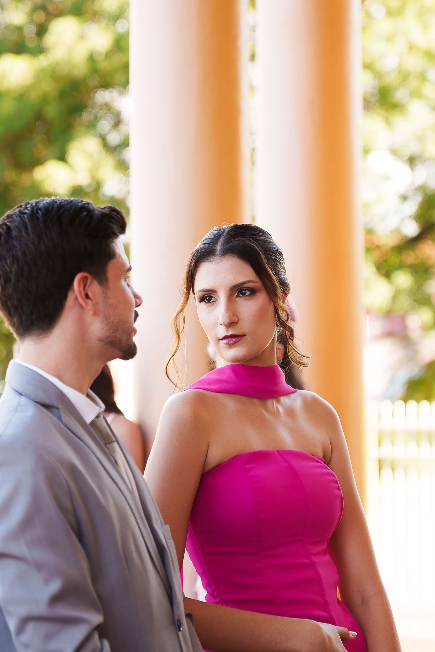 Elegant couple in formal attire, woman in pink halter dress gazes at camera, man in gray suit, outdoor columns backdrop.