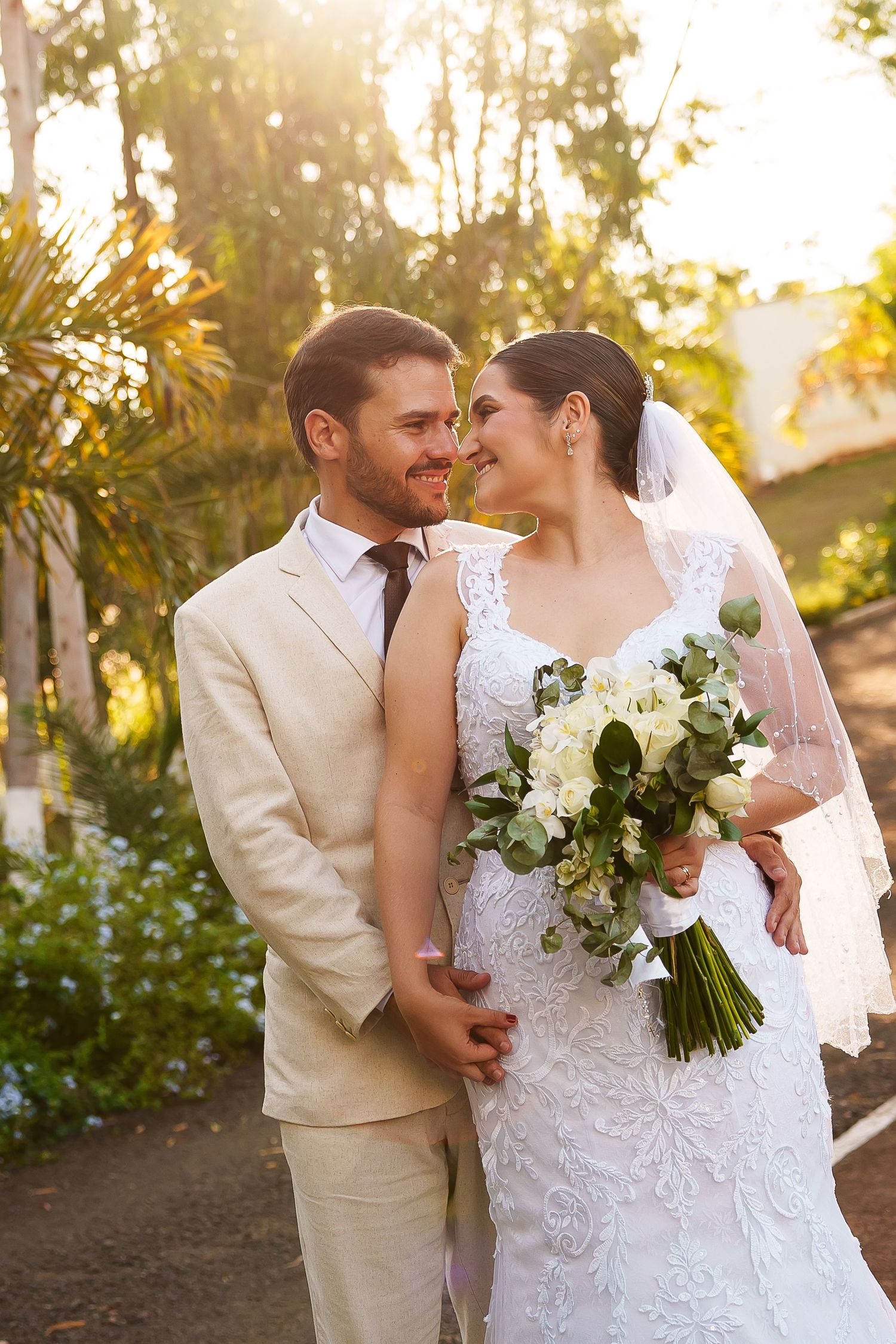 A bride and groom share a romantic moment outdoors, with the bride holding a white floral bouquet in golden sunlight.