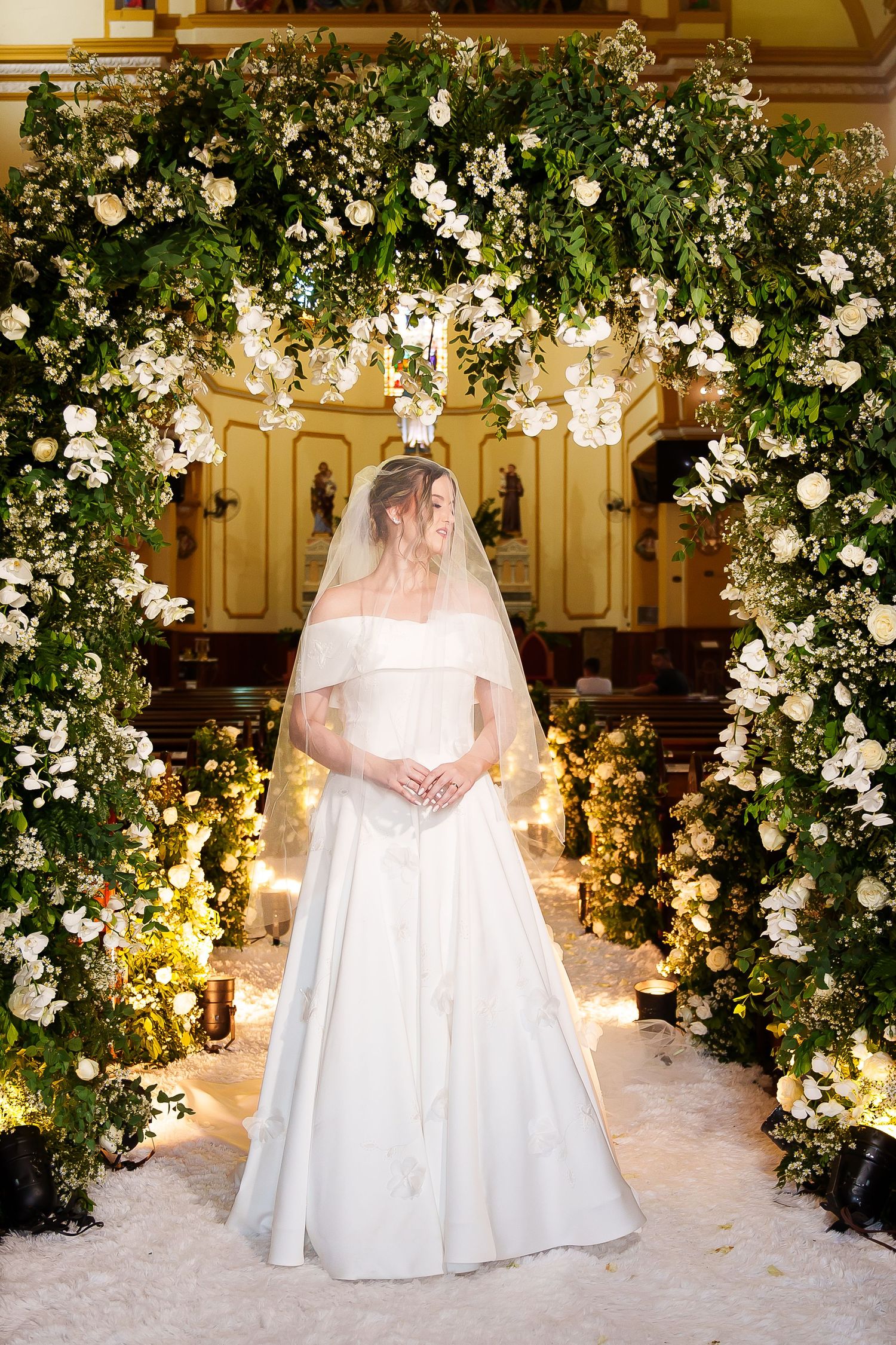 Bride in white off-shoulder gown and veil stands under lush floral arch with white roses in elegant church.
