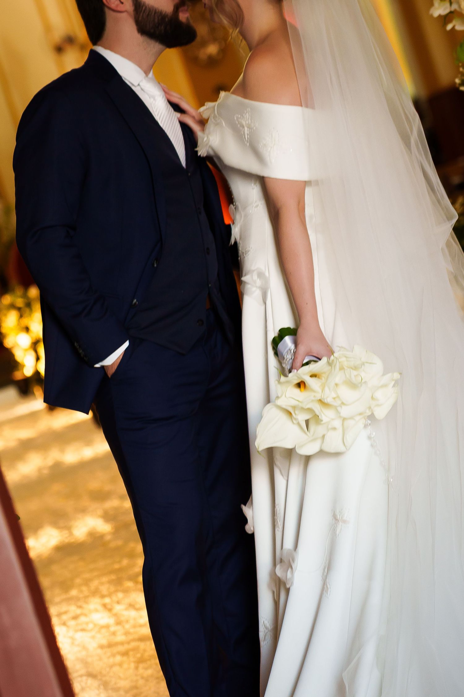 Bride in white off-shoulder gown holding calla lily bouquet stands beside groom in navy suit, sharing a kiss.