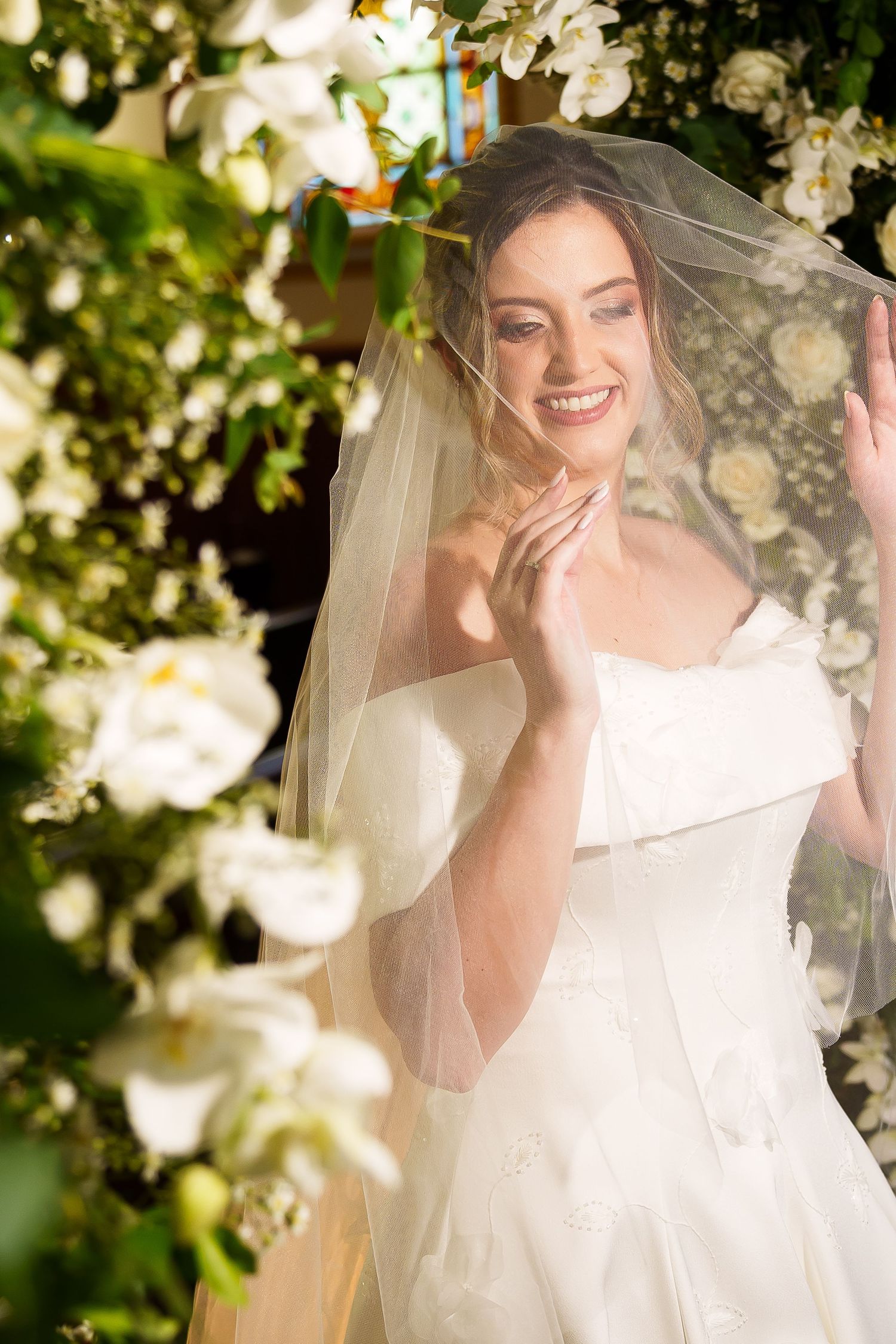Smiling bride in white gown and veil surrounded by lush white floral arrangements on her wedding day.