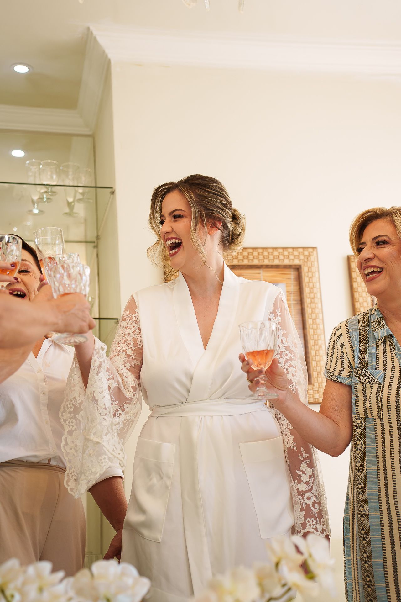 Bride in white lace robe laughing with bridesmaids holding champagne glasses during wedding morning preparations.