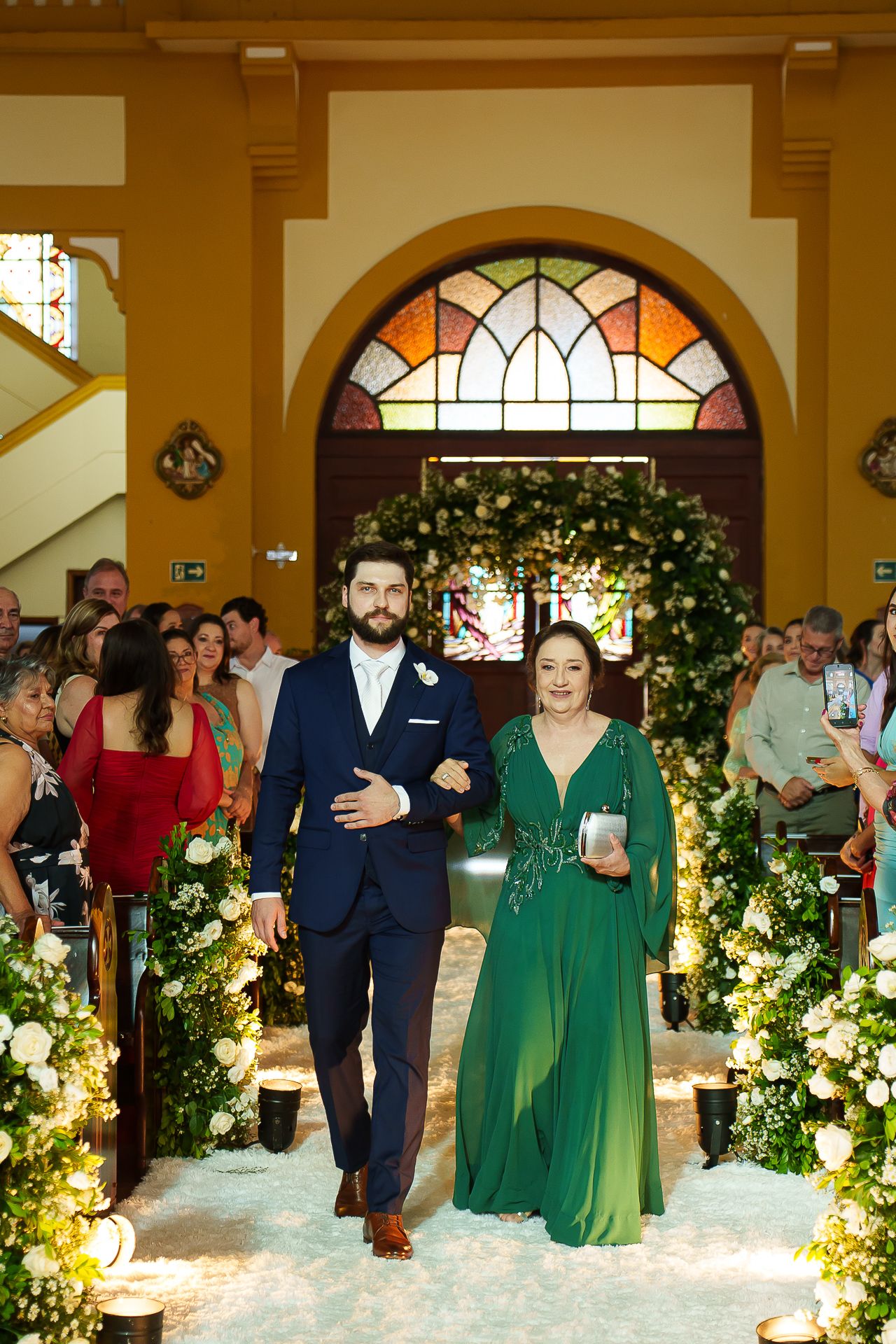 Groom in navy suit walks bride's mother in green gown down floral-lined church aisle during wedding ceremony.