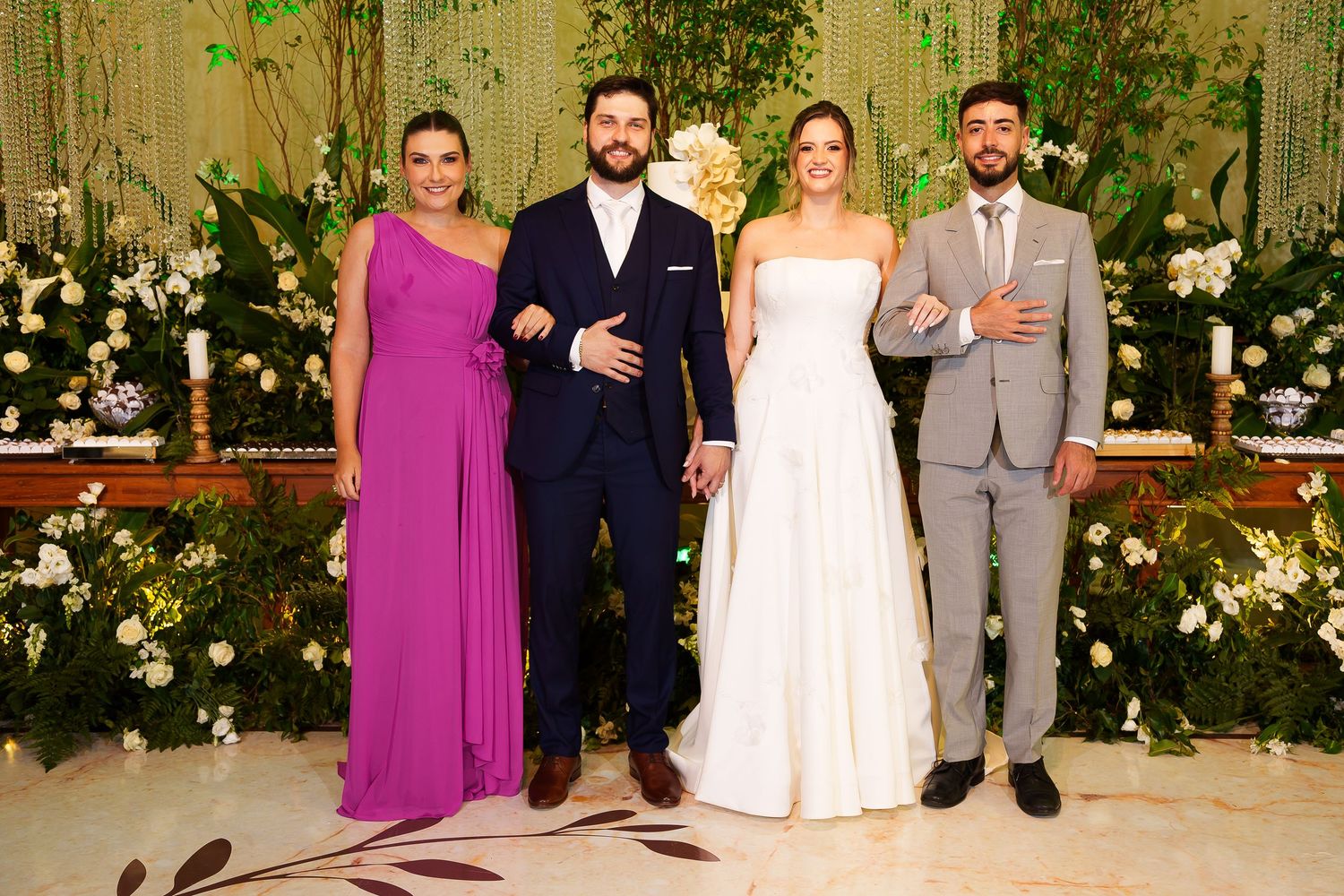 Wedding party of four posing at floral altar, bride in white gown, groom in navy suit, guests in purple dress and gray suit.