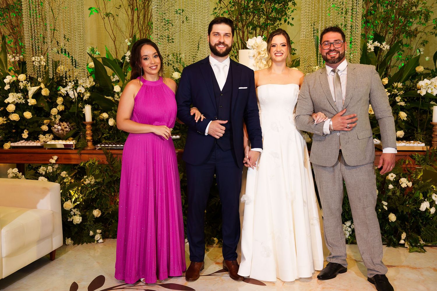Bride and groom pose with two guests at elegant wedding reception with lush green floral backdrop.