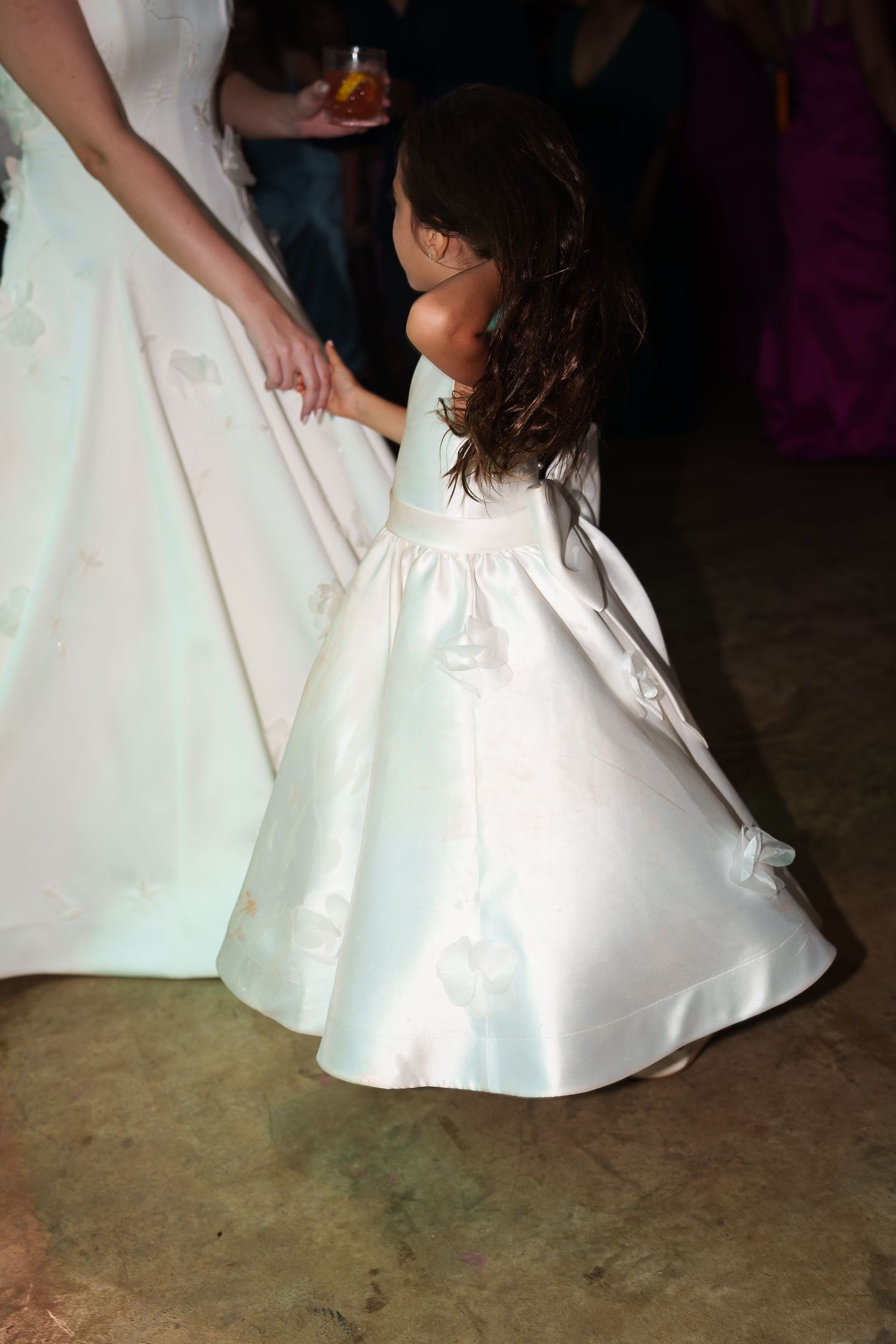 Young flower girl in white satin gown holds bride's dress at wedding reception.