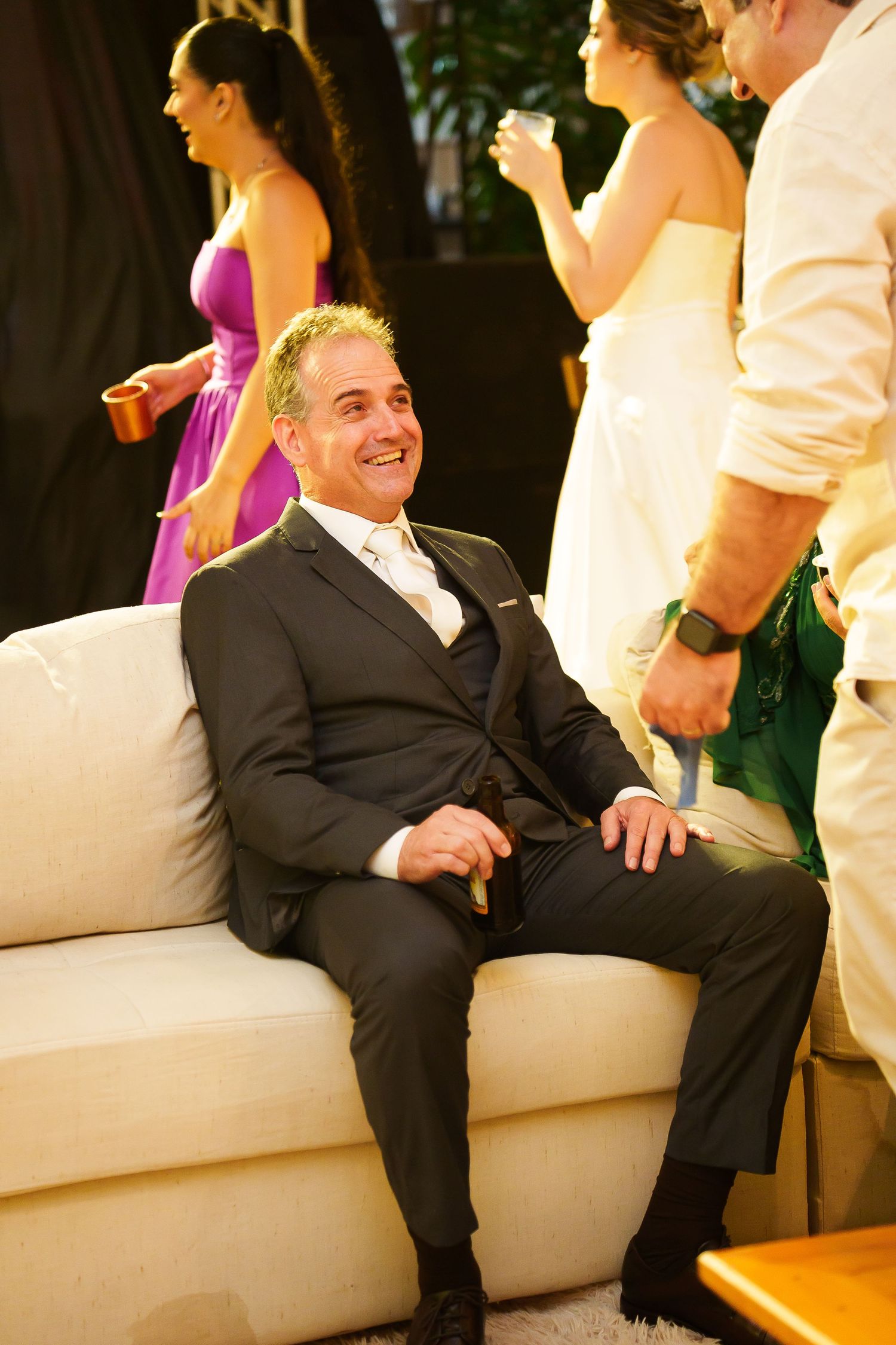 Smiling man in dark suit relaxing on white sofa at elegant wedding reception event.