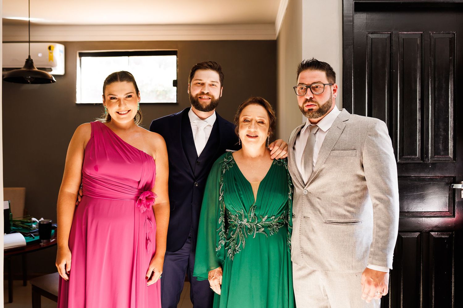 Four people posing indoors, wearing formal attire including pink, green, and beige outfits, smiling for a group photo.