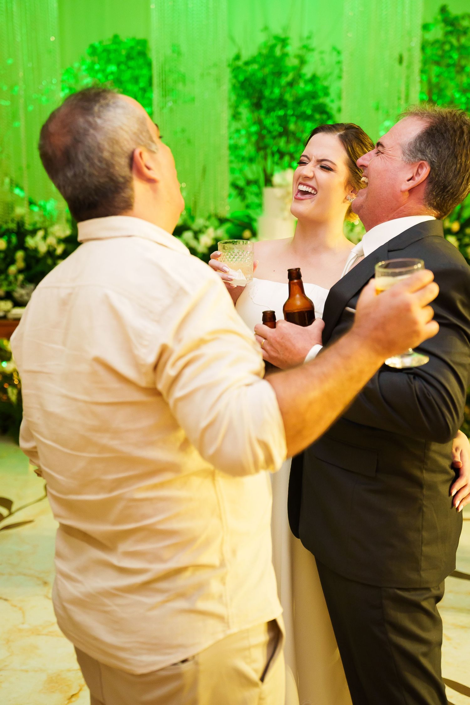 Bride and groom laughing with guest at wedding reception, holding drinks with green floral backdrop.