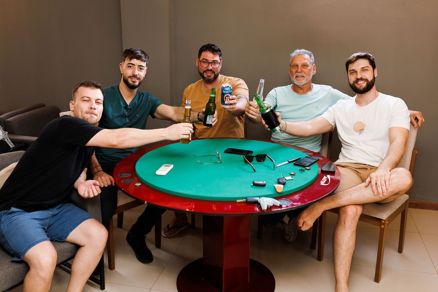 Five men smiling and raising drinks around a green poker table in a casual indoor setting.