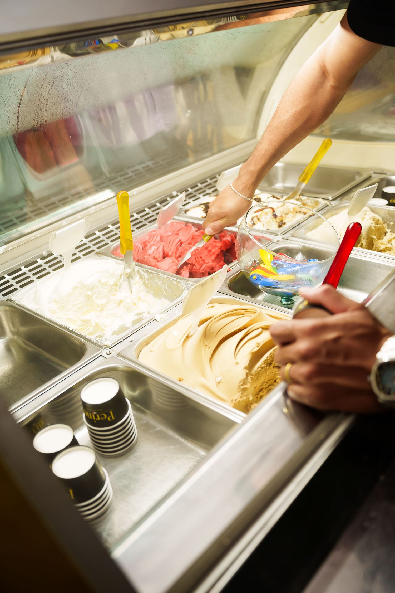 Person serving colorful gelato flavors from a stainless steel display case in an ice cream shop.