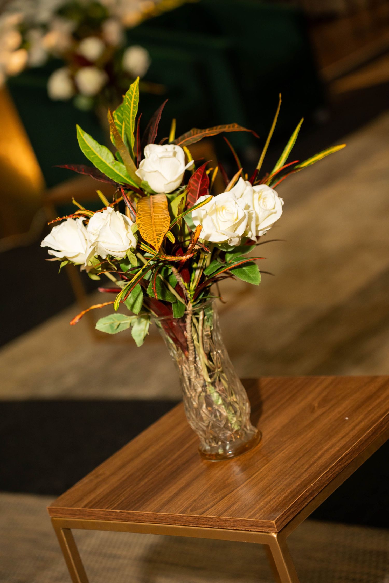 Crystal vase with white roses and autumn foliage on a wooden table in warm golden light.