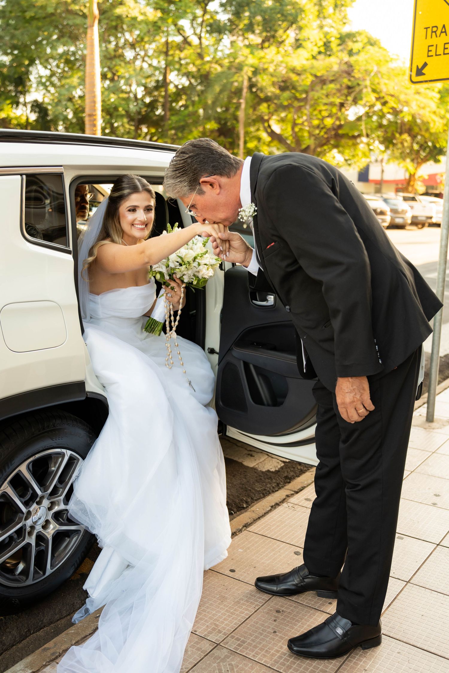 A groom in a black suit kisses the hand of a bride in a white gown as she exits a white vehicle holding a bouquet.