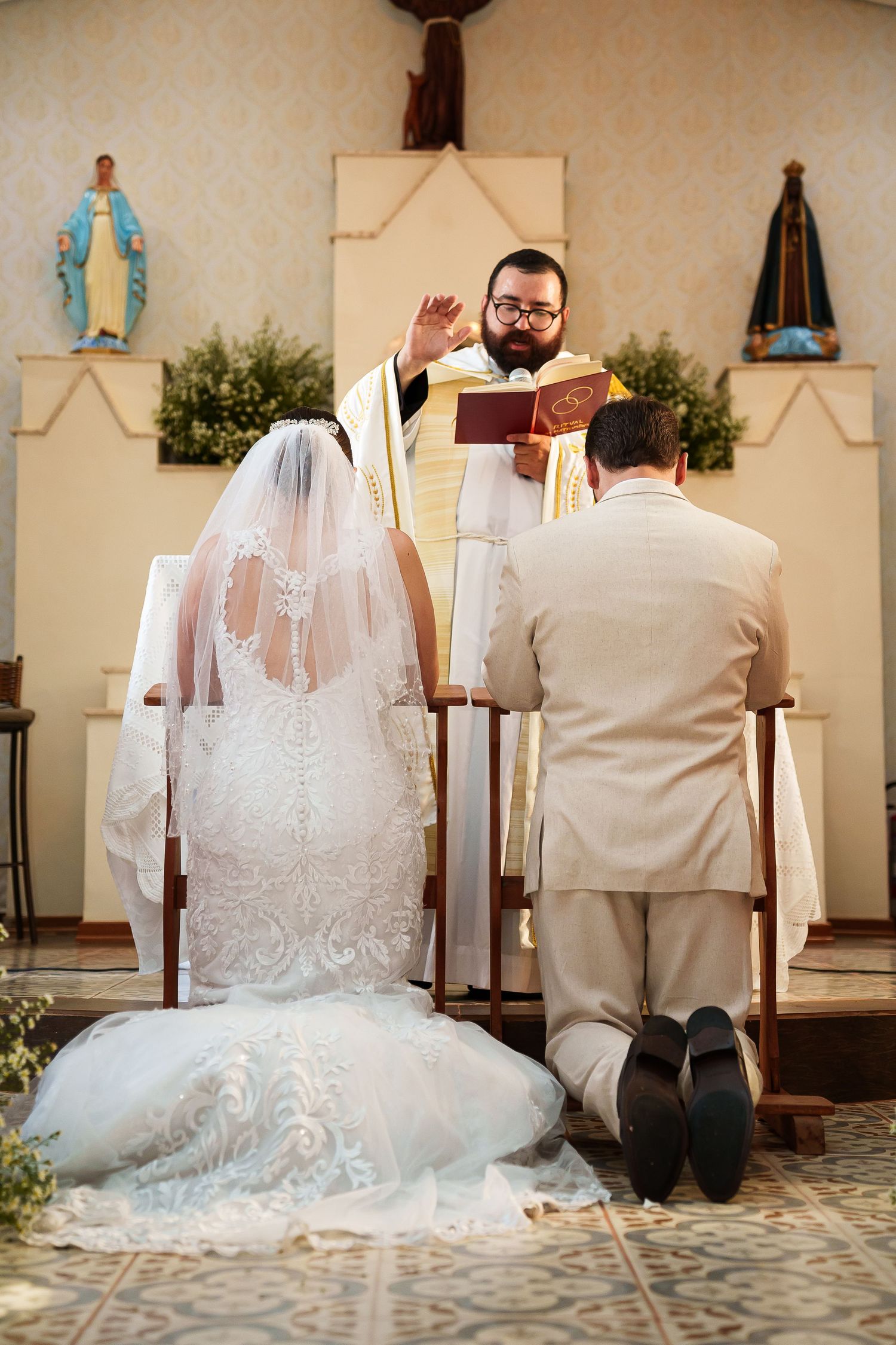 Bride and groom kneeling at altar during Catholic wedding ceremony as priest performs blessing in church.