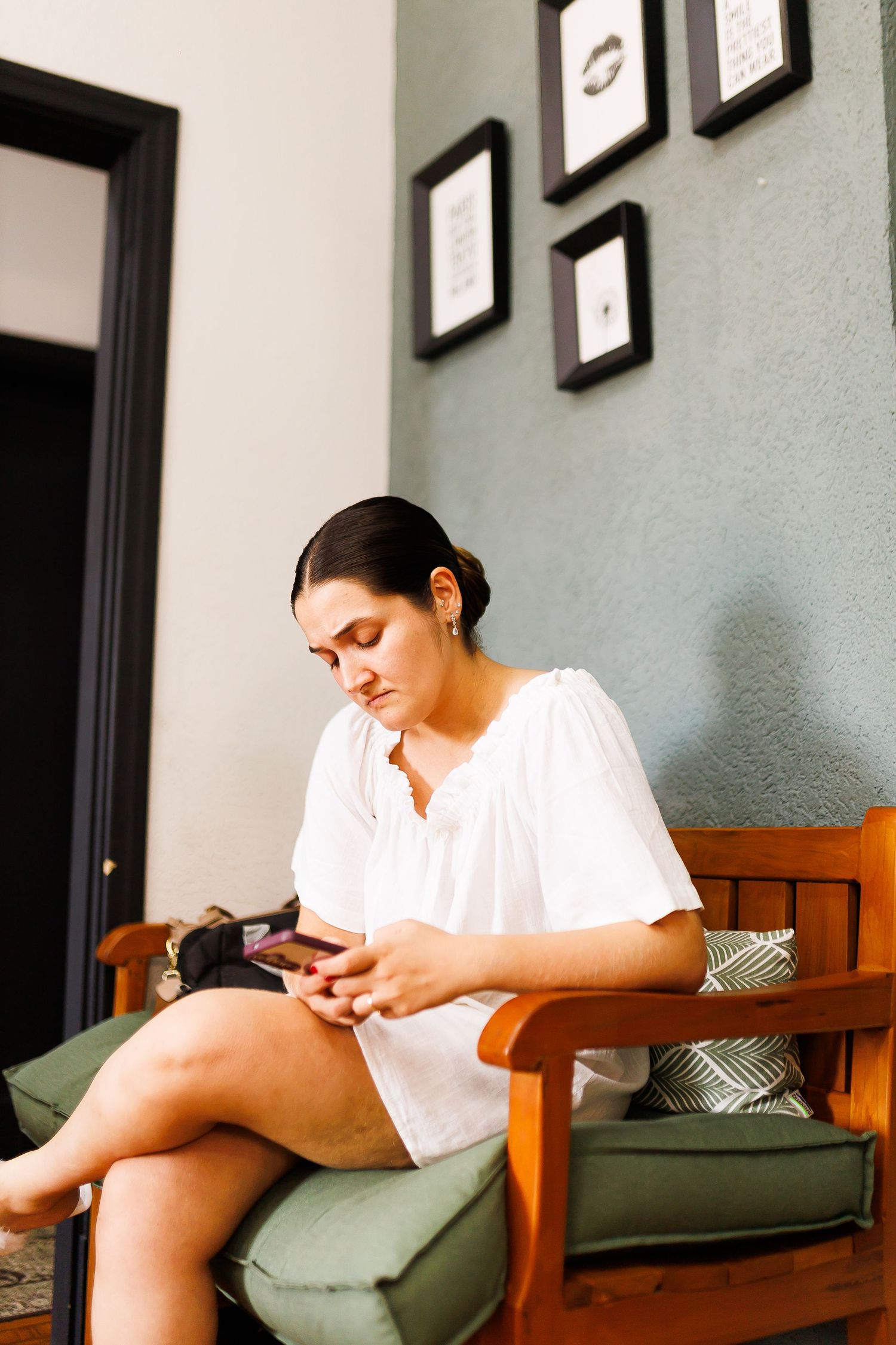 Woman in white blouse sitting on wooden bench using smartphone, with framed wall art in background.