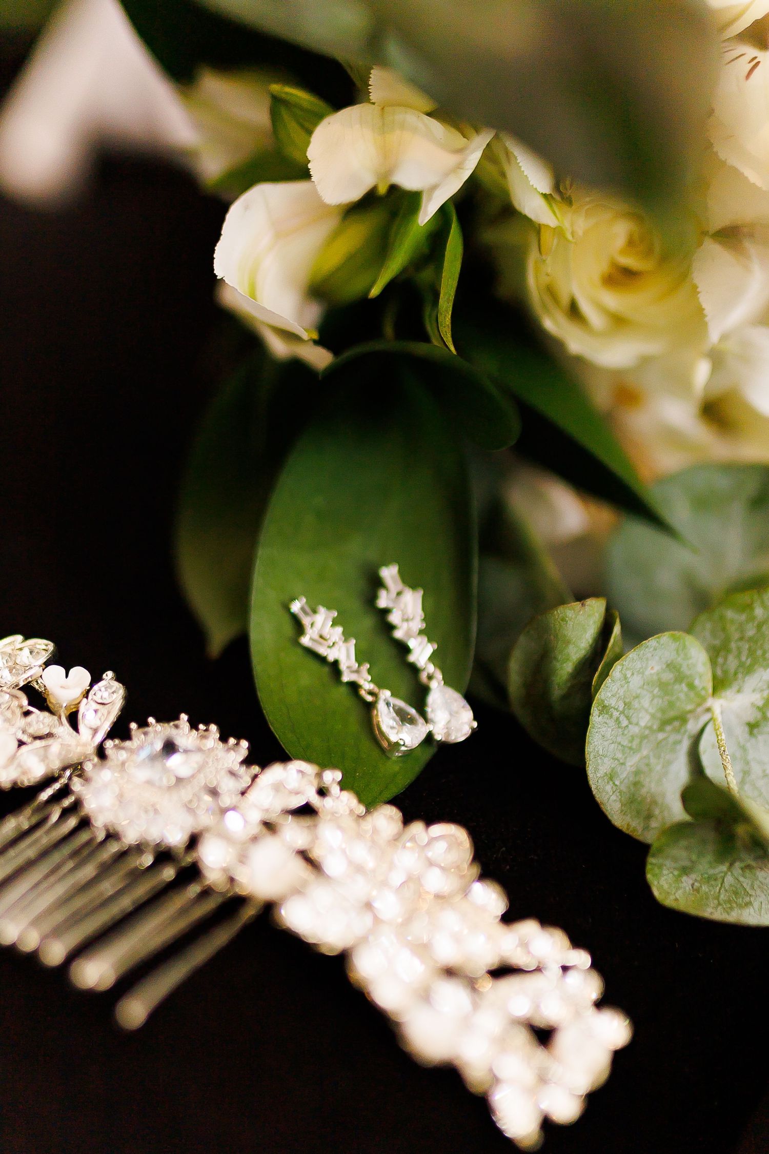 Elegant bridal accessories featuring crystal drop earrings, ornate silver hair comb, and white floral bouquet with eucalyptus greenery.