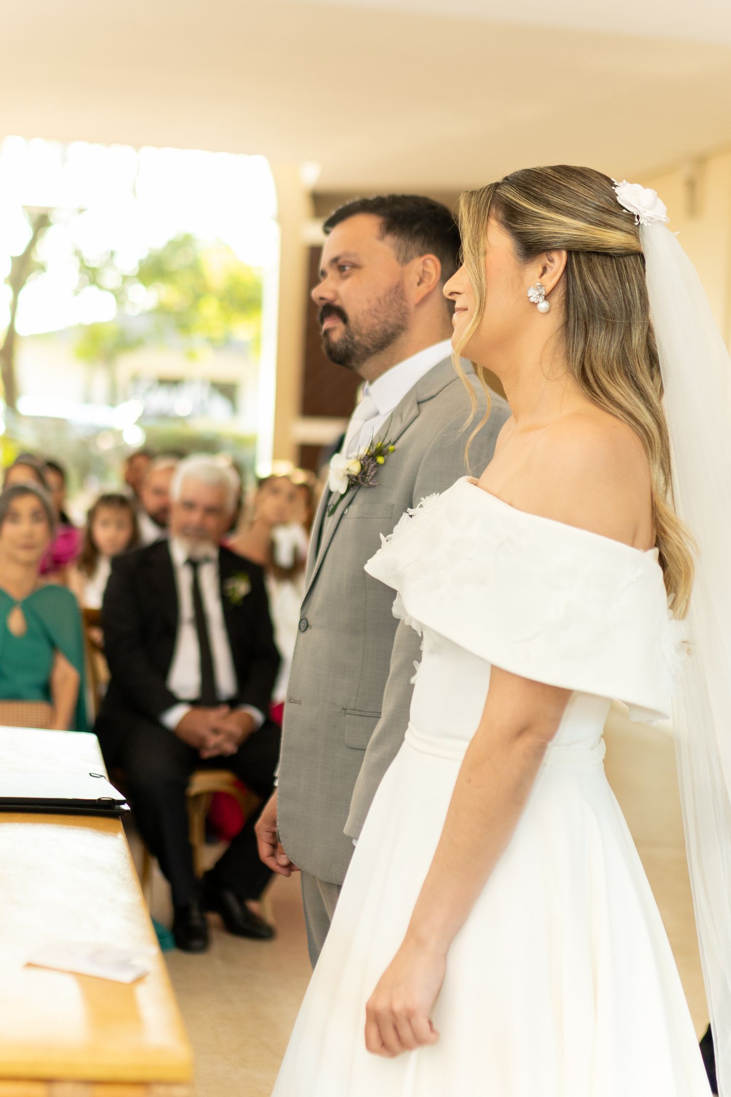 Bride in white off-shoulder gown and groom in gray suit stand at altar during wedding ceremony.