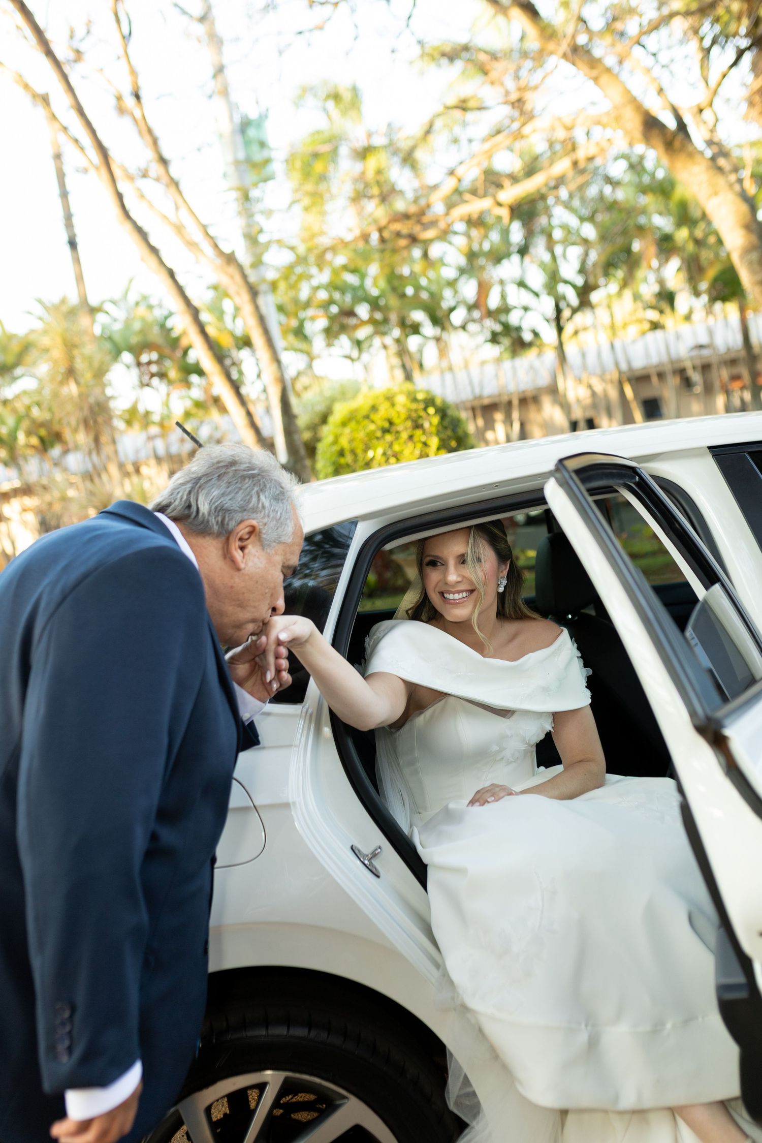 Bride in white off-shoulder gown exits white limo as man in navy suit kisses her hand outdoors.