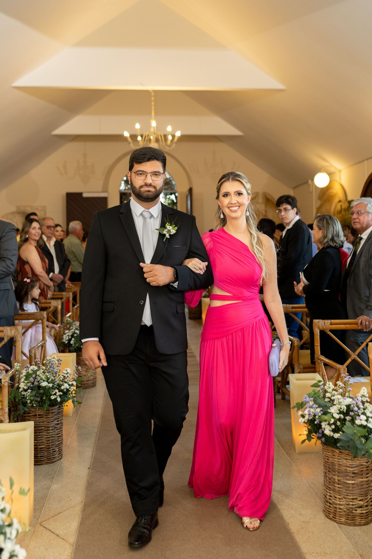 Couple walks down wedding aisle, man in black suit, woman in pink one-shoulder gown, chapel setting.