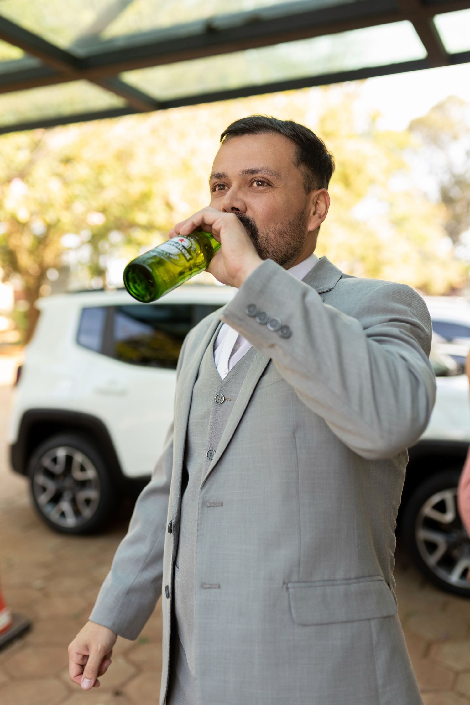 Man in gray suit drinking from green bottle near white SUV outdoors.
