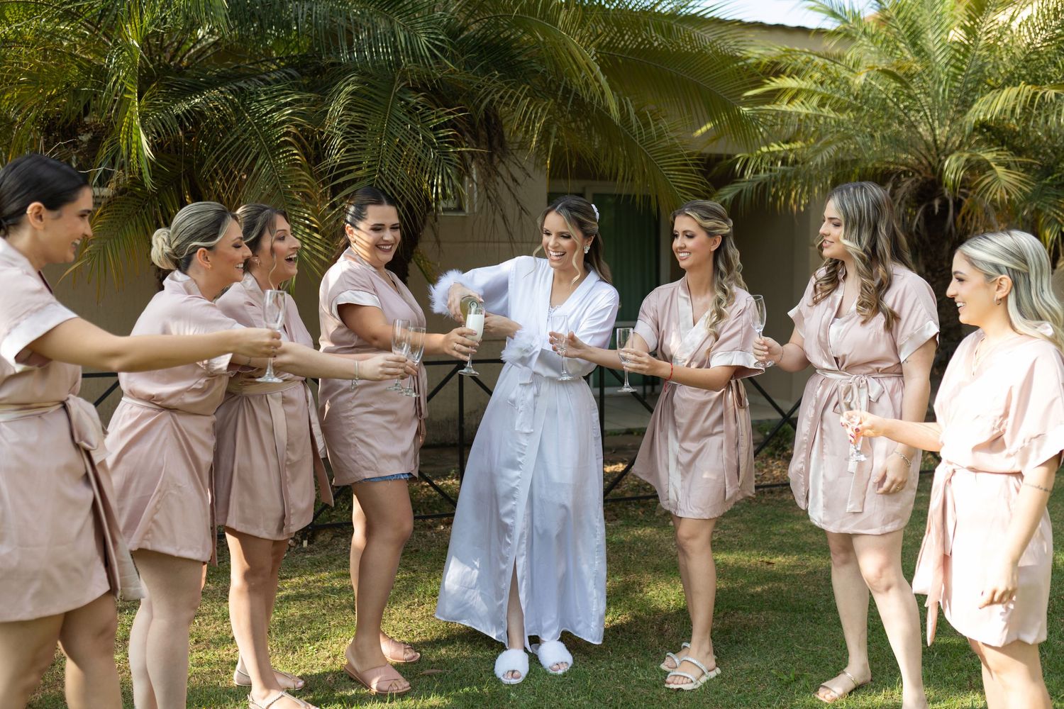 Bride in white robe celebrates with bridesmaids in tan robes, toasting champagne outdoors near tropical palm trees.