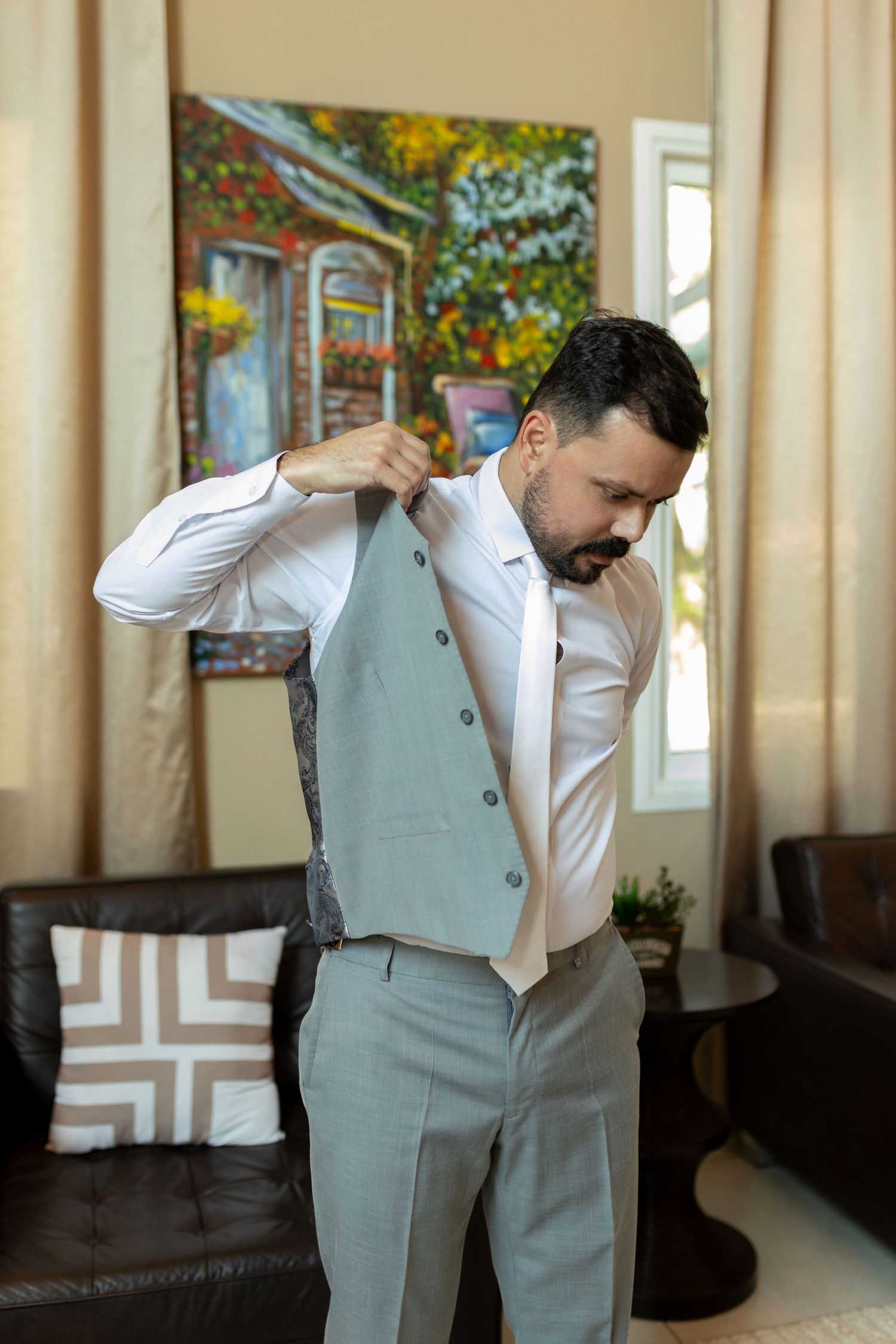 Man in gray vest and white shirt adjusting his tie while getting dressed in a well-decorated living room.