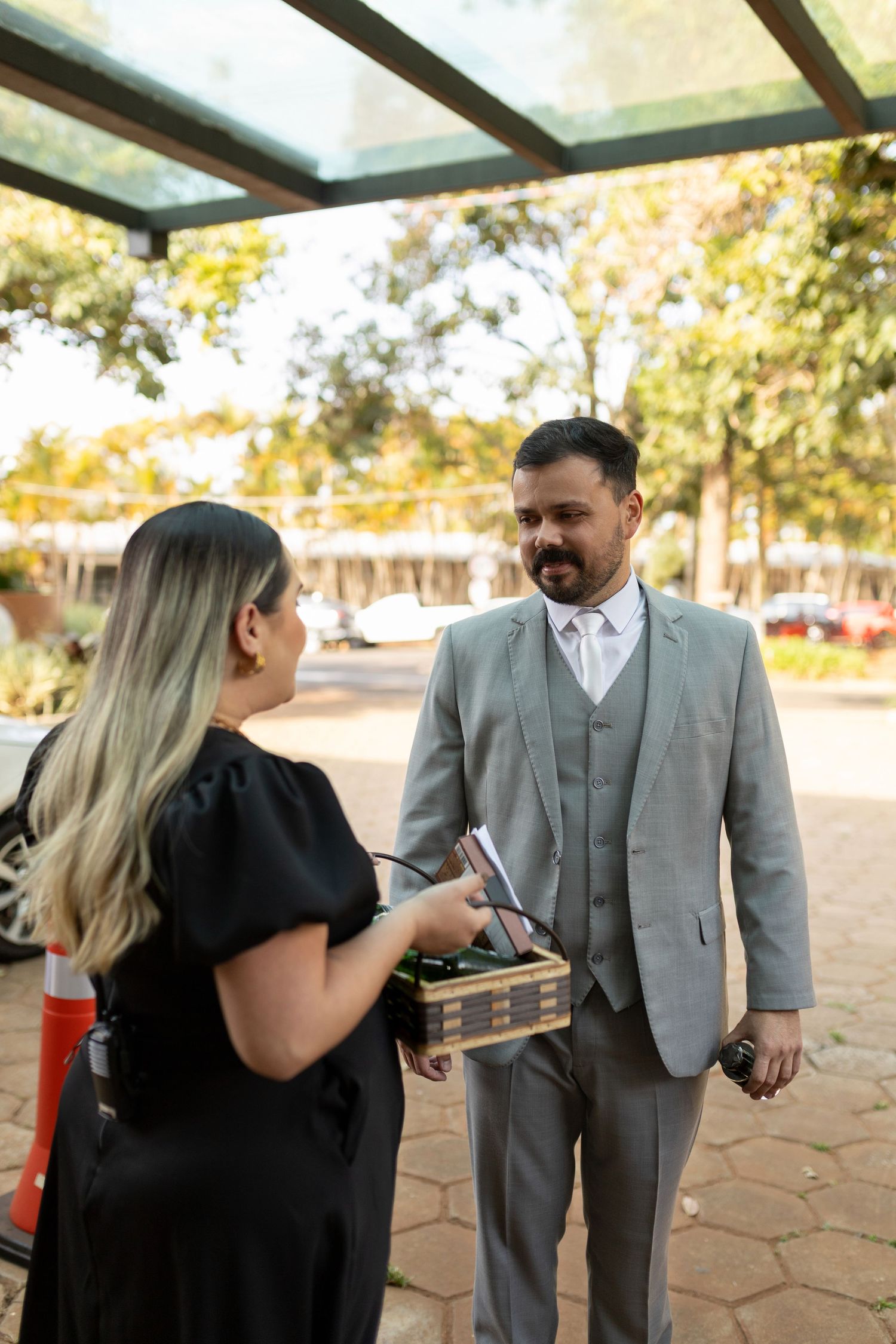 Man in gray suit receiving a wooden box from a woman in black dress outdoors under a glass canopy.
