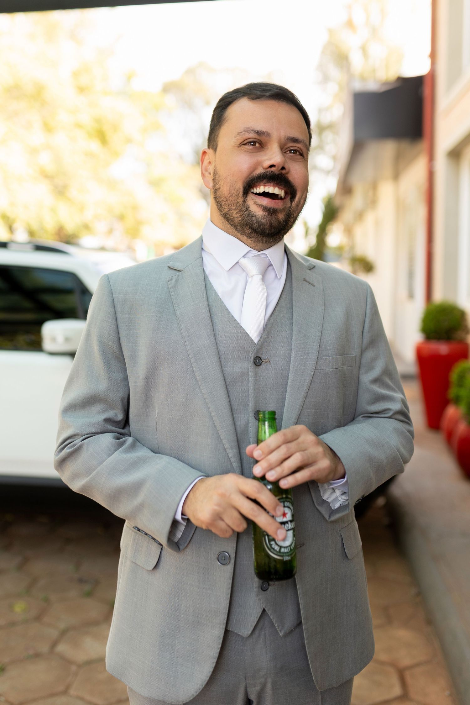 Smiling man in gray suit holding green beer bottle outdoors near white car and red planter.