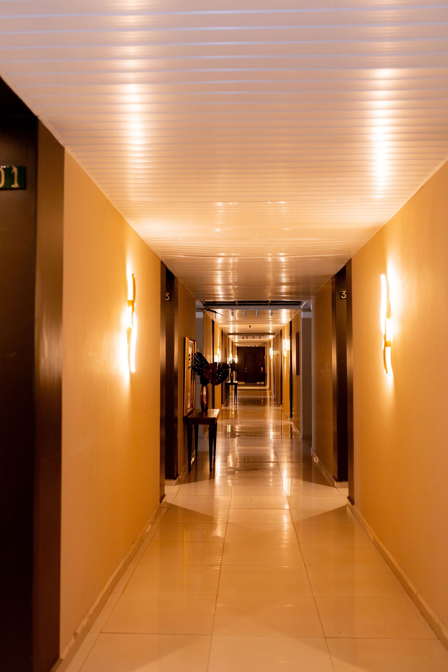 Elegant hotel corridor with warm golden walls, wall sconces, and reflective tiled floors leading to a vanishing point.