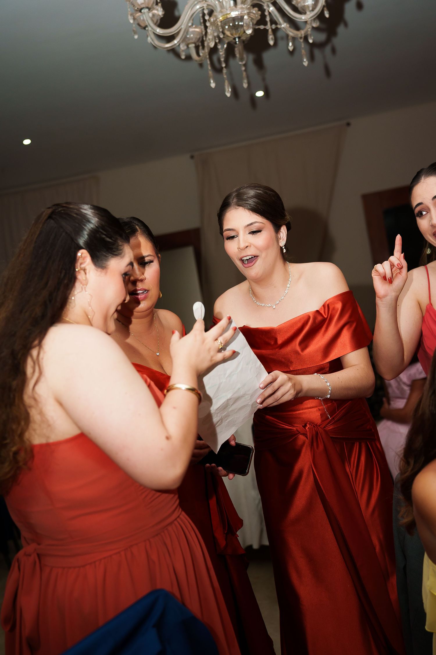 Bridesmaids in red satin gowns gather excitedly reading a note under a crystal chandelier at a wedding celebration.