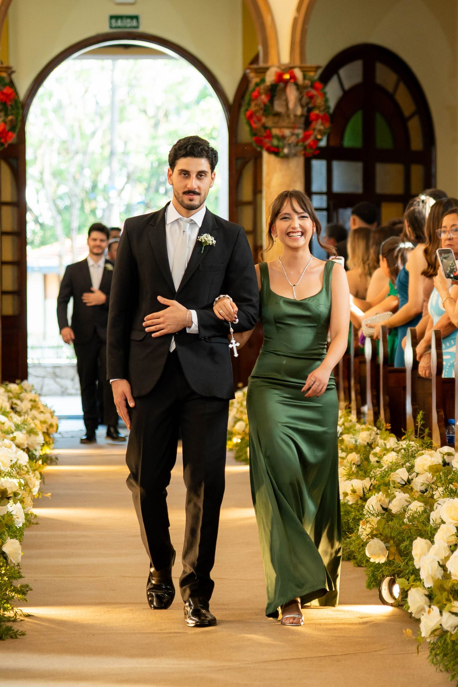Elegant couple walking down a flower-lined church aisle, man in black tuxedo and woman in green satin gown.