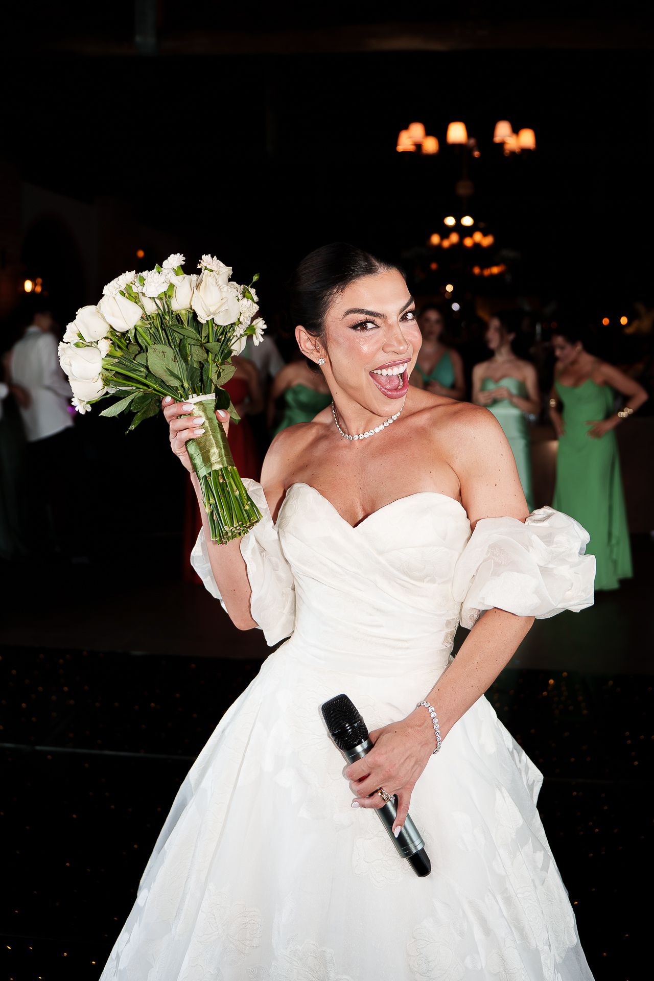 Excited bride in white off-shoulder gown holds bouquet and microphone, laughing joyfully at wedding reception.