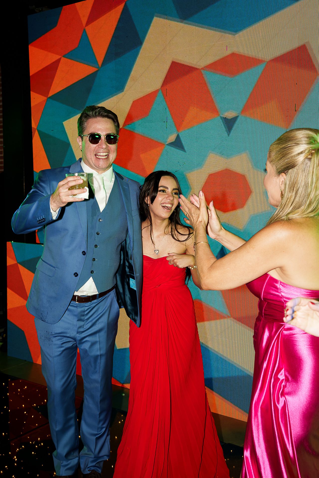 Three guests in formal attire dance at a colorful event, man in blue suit raising a glass with women in red and pink gowns.