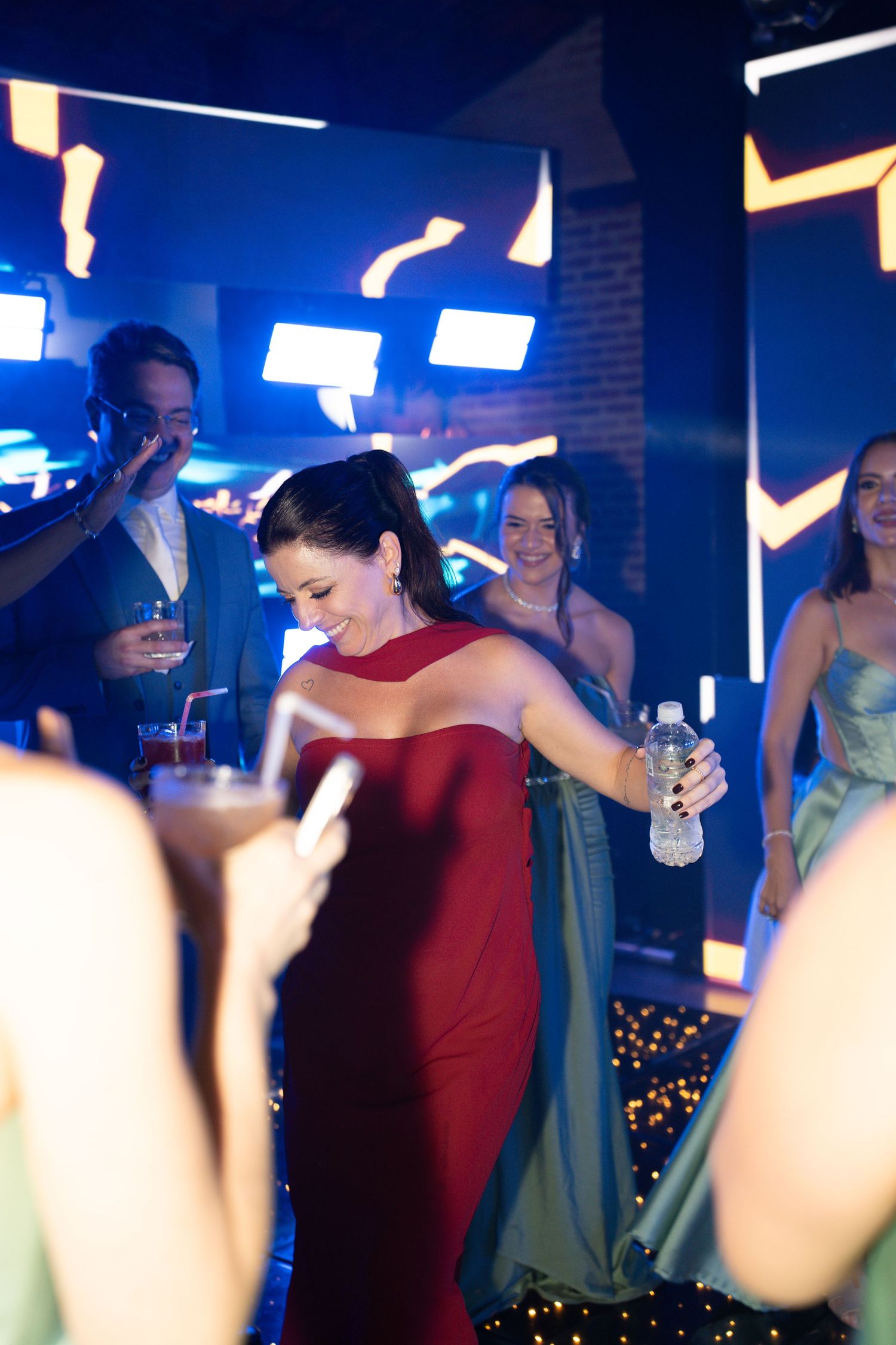 Young woman in red gown dancing at prom party with friends in formal wear under blue neon lights.