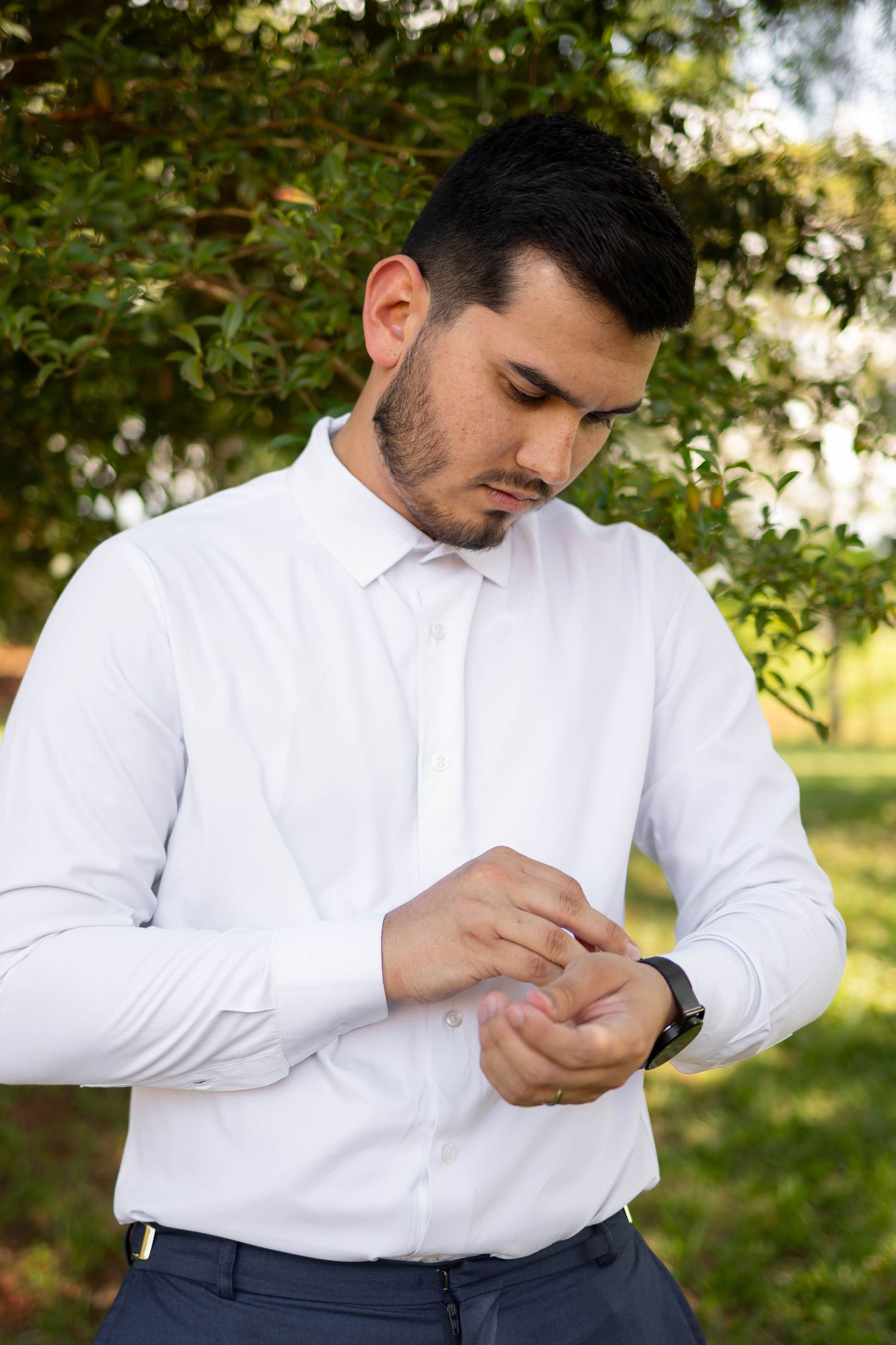 Young man in white dress shirt adjusting cufflinks outdoors with green trees in background.