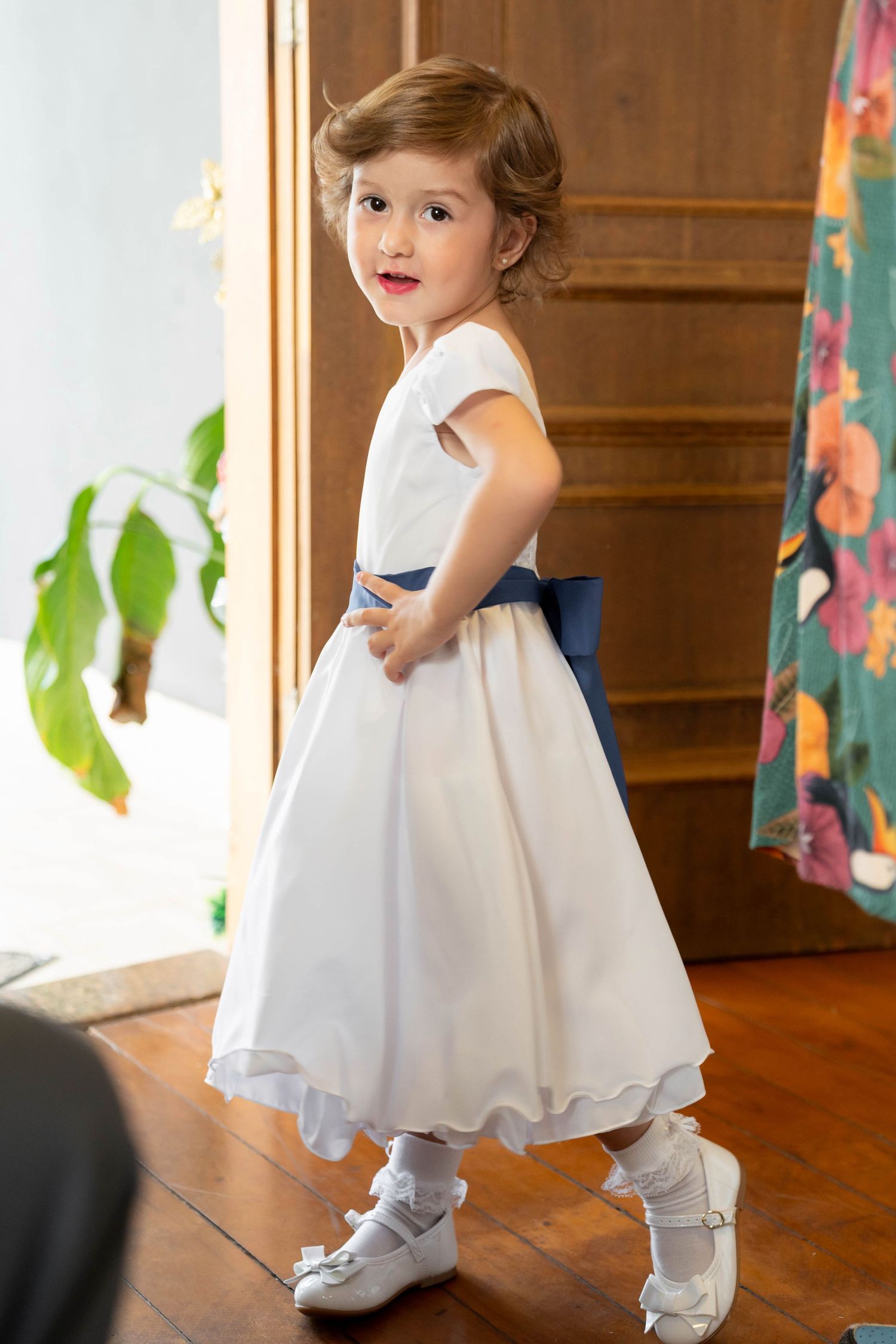 Young girl in white dress with blue sash bow, white shoes and socks, posing indoors on hardwood floor.