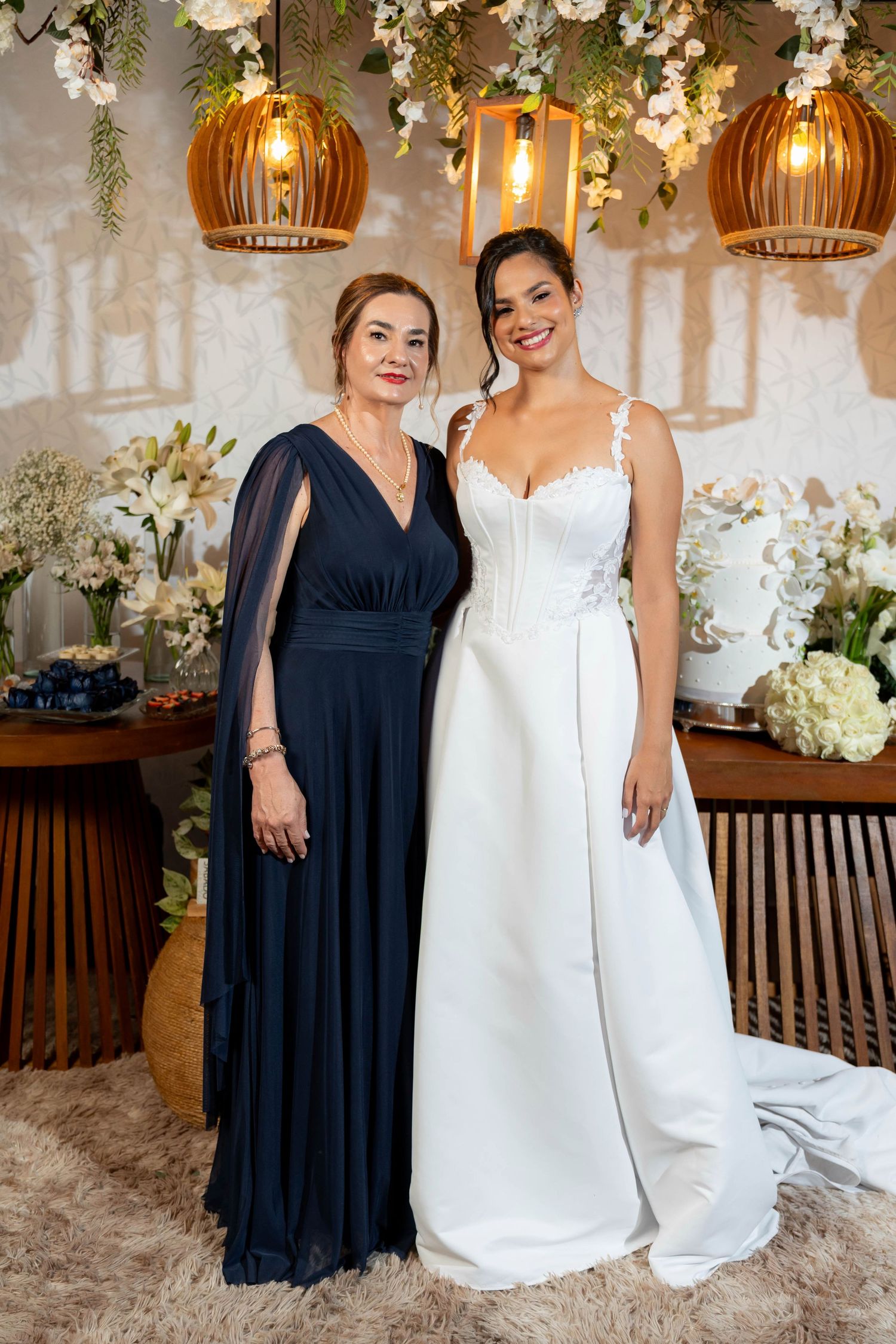 Bride in white gown poses with woman in navy blue dress at elegant wedding reception with floral decor.