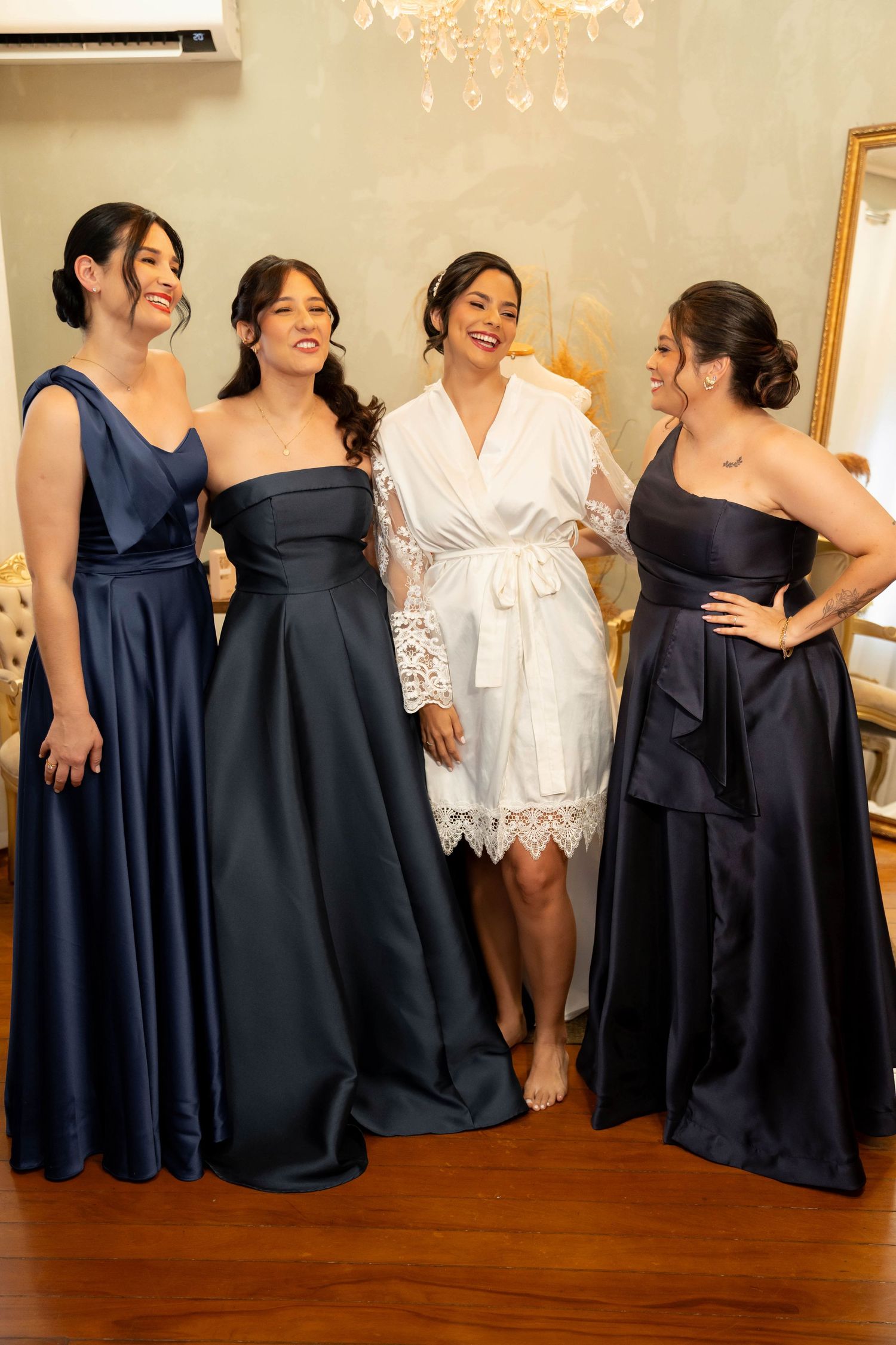 Bride in white lace robe poses with three bridesmaids in navy blue gowns in an elegant room.