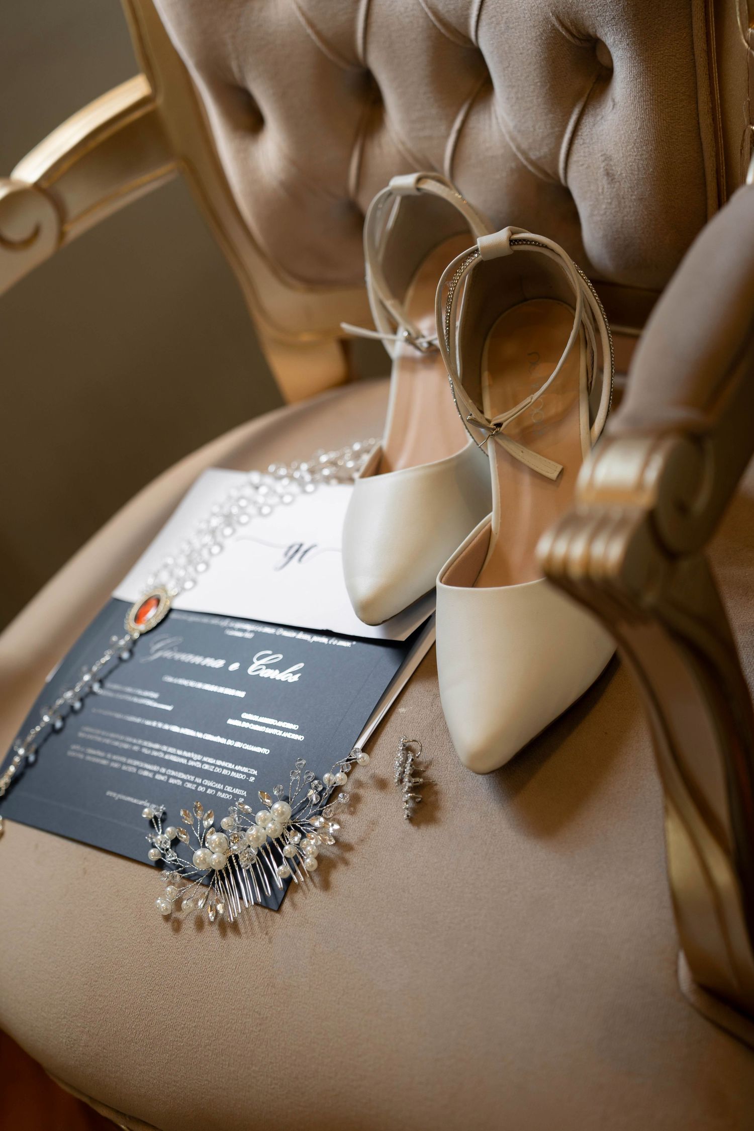 Elegant bridal flat lay featuring ivory heels, pearl hair accessories, and wedding stationery on a tufted beige chair.