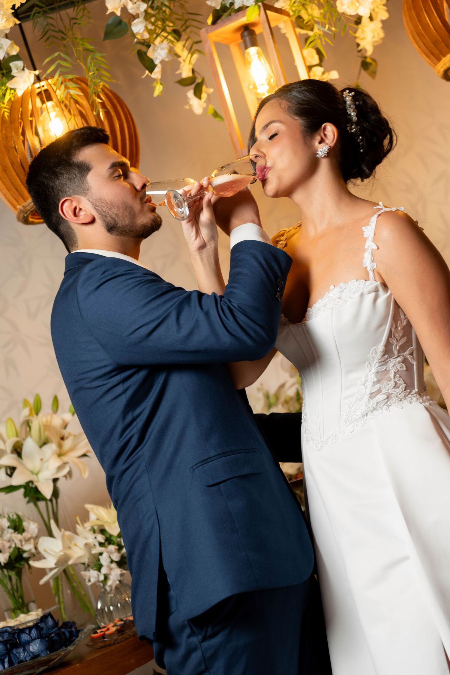Bride and groom sharing a champagne toast at their wedding reception surrounded by white flowers and warm lighting.