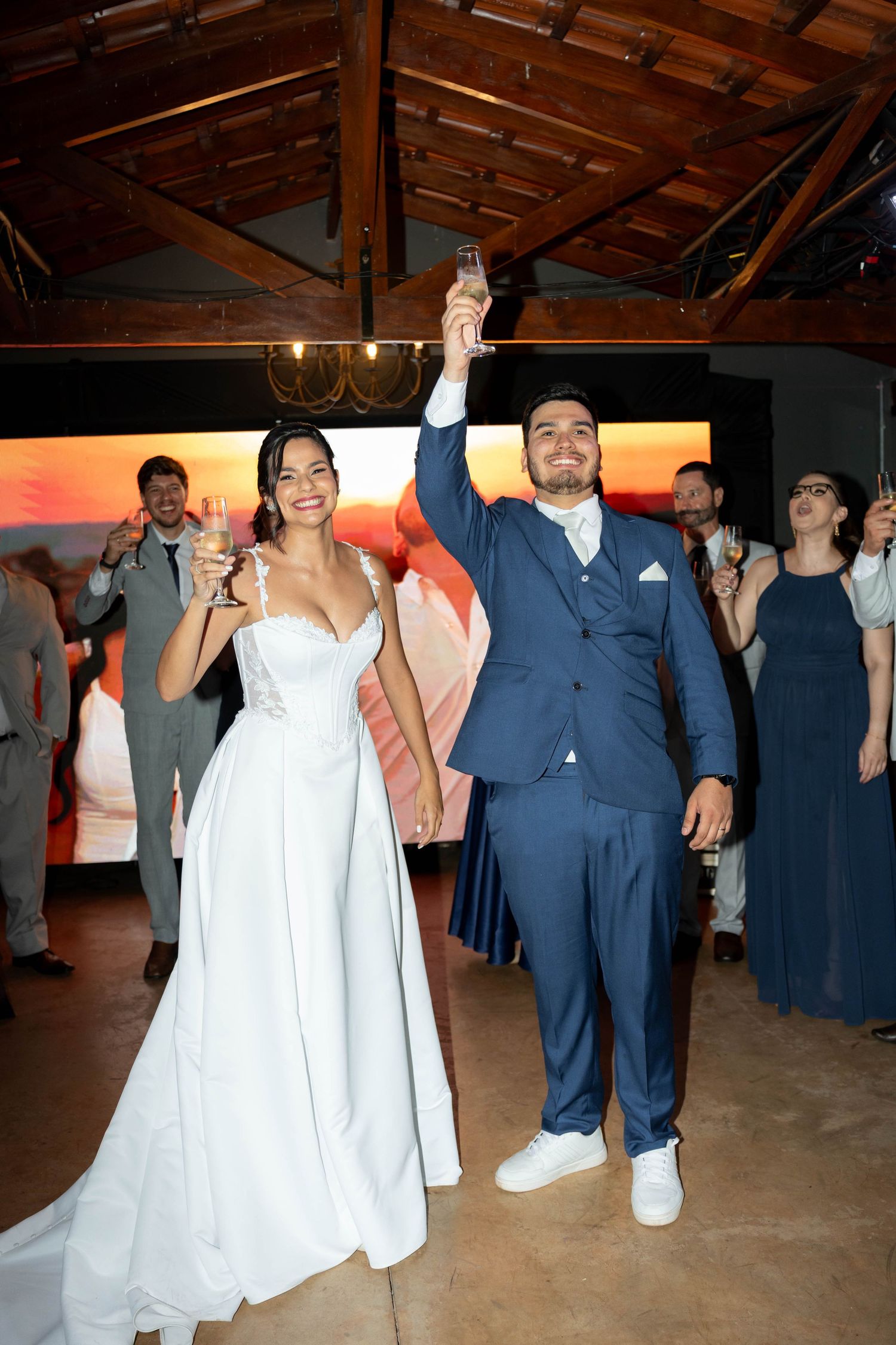 Bride in white gown and groom in blue suit raising champagne glasses in celebratory toast at wedding reception.
