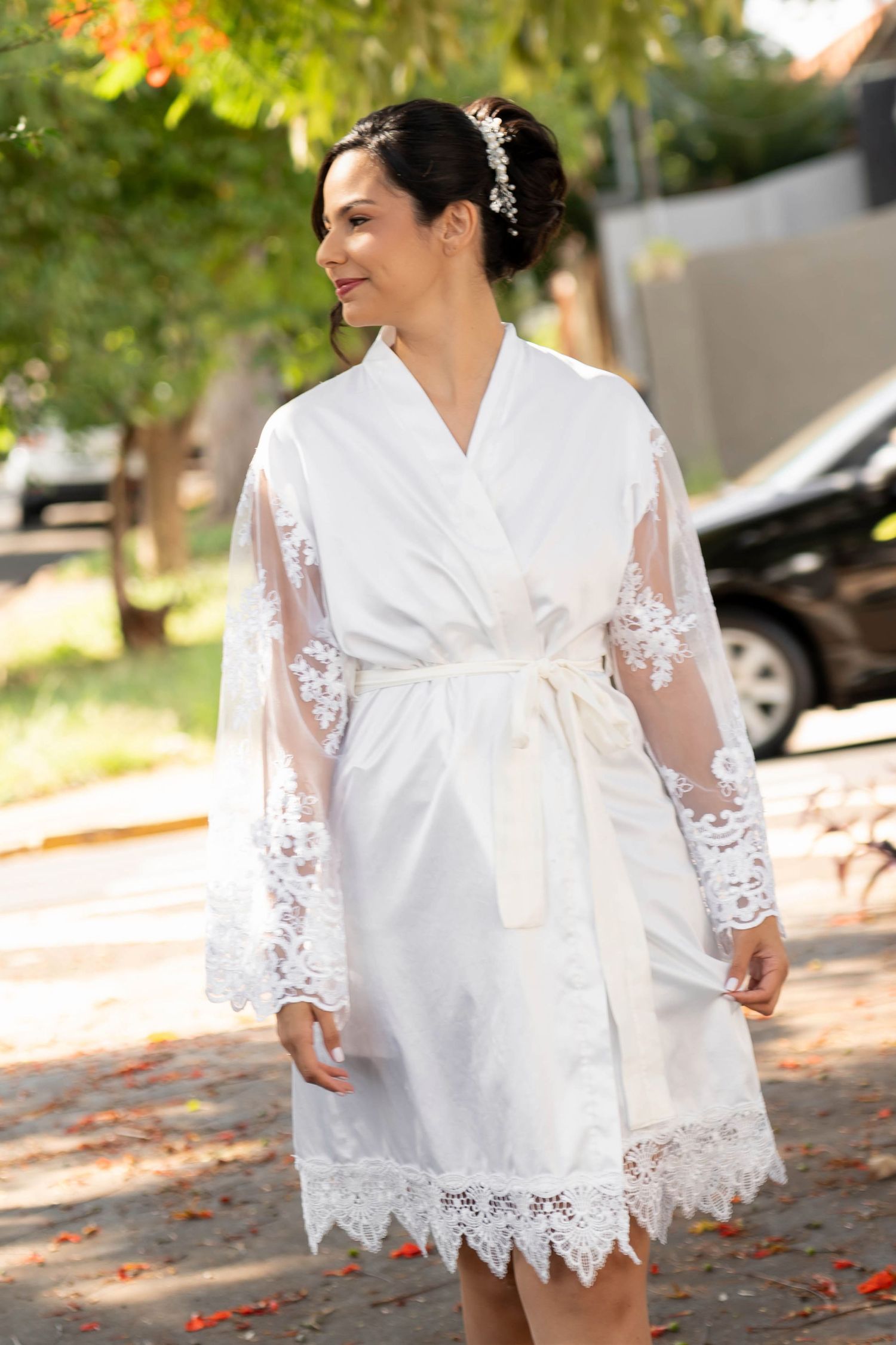 Smiling bride in white lace robe with sheer sleeves and floral hair accessory standing outdoors.