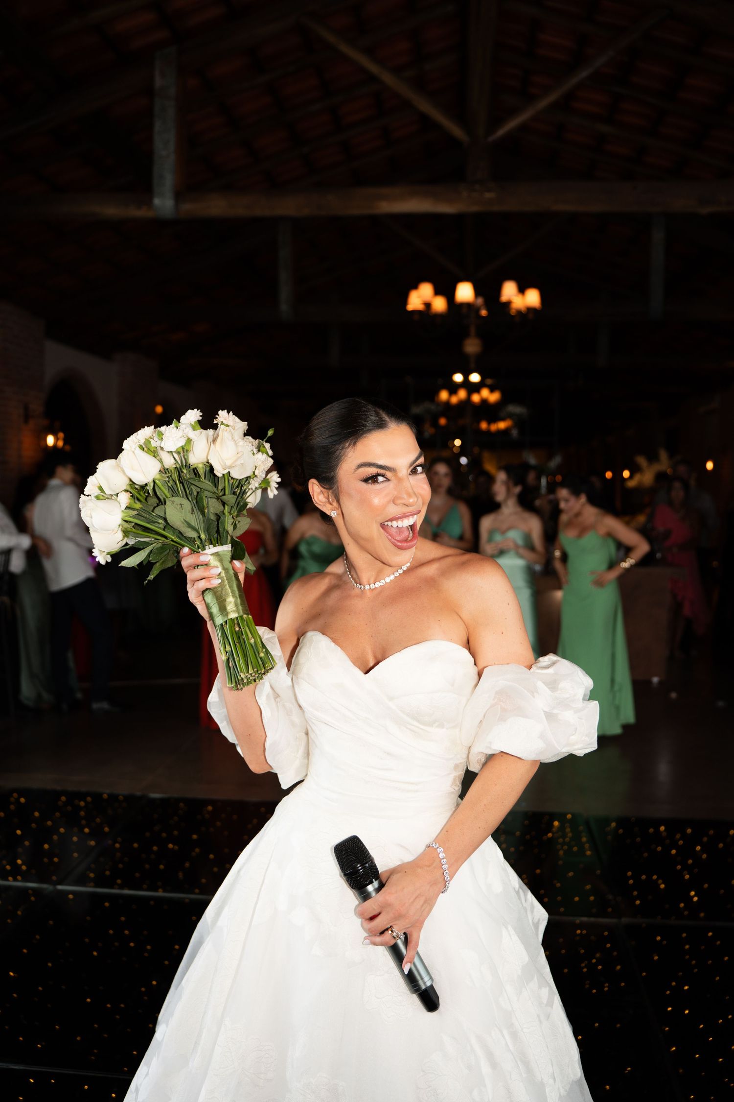 Bride in white ball gown holds bouquet and microphone, laughing joyfully on dance floor at elegant wedding reception.
