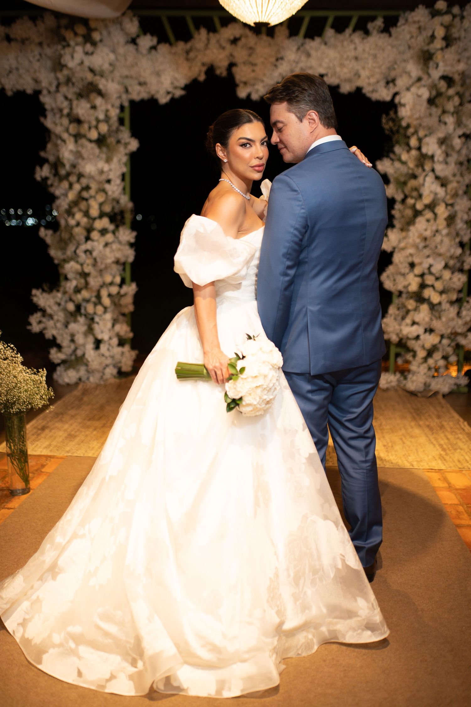 Bride in white off-shoulder ball gown holding bouquet poses with groom in blue suit before floral arch.