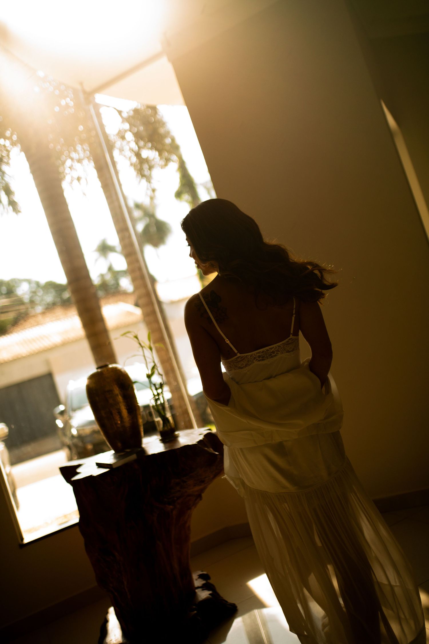 Woman in white dress silhouetted against warm sunlight streaming through large windows with tropical palm trees outside.