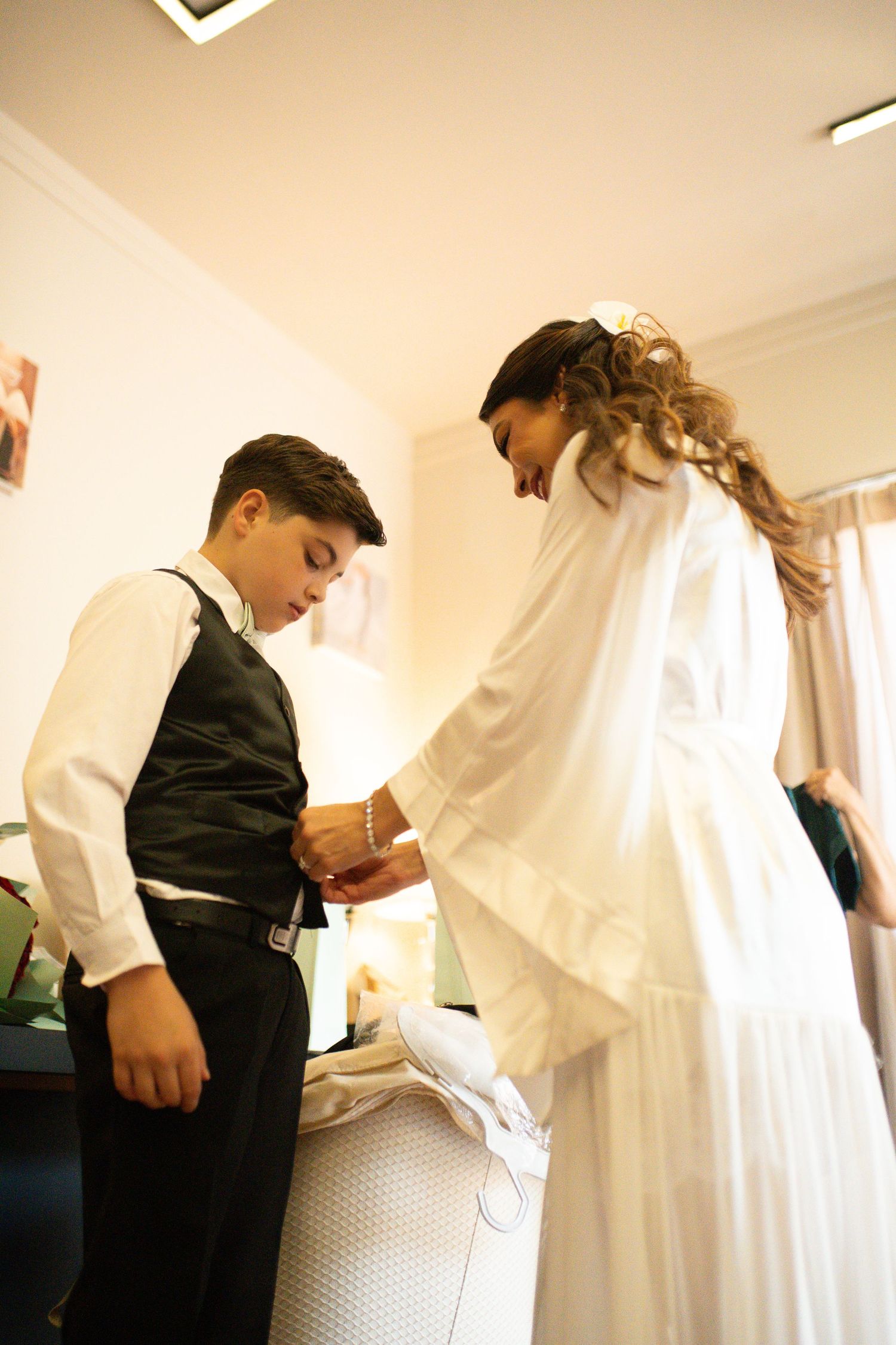 Bride in white gown helping young boy in black vest adjust his cufflinks before wedding ceremony.