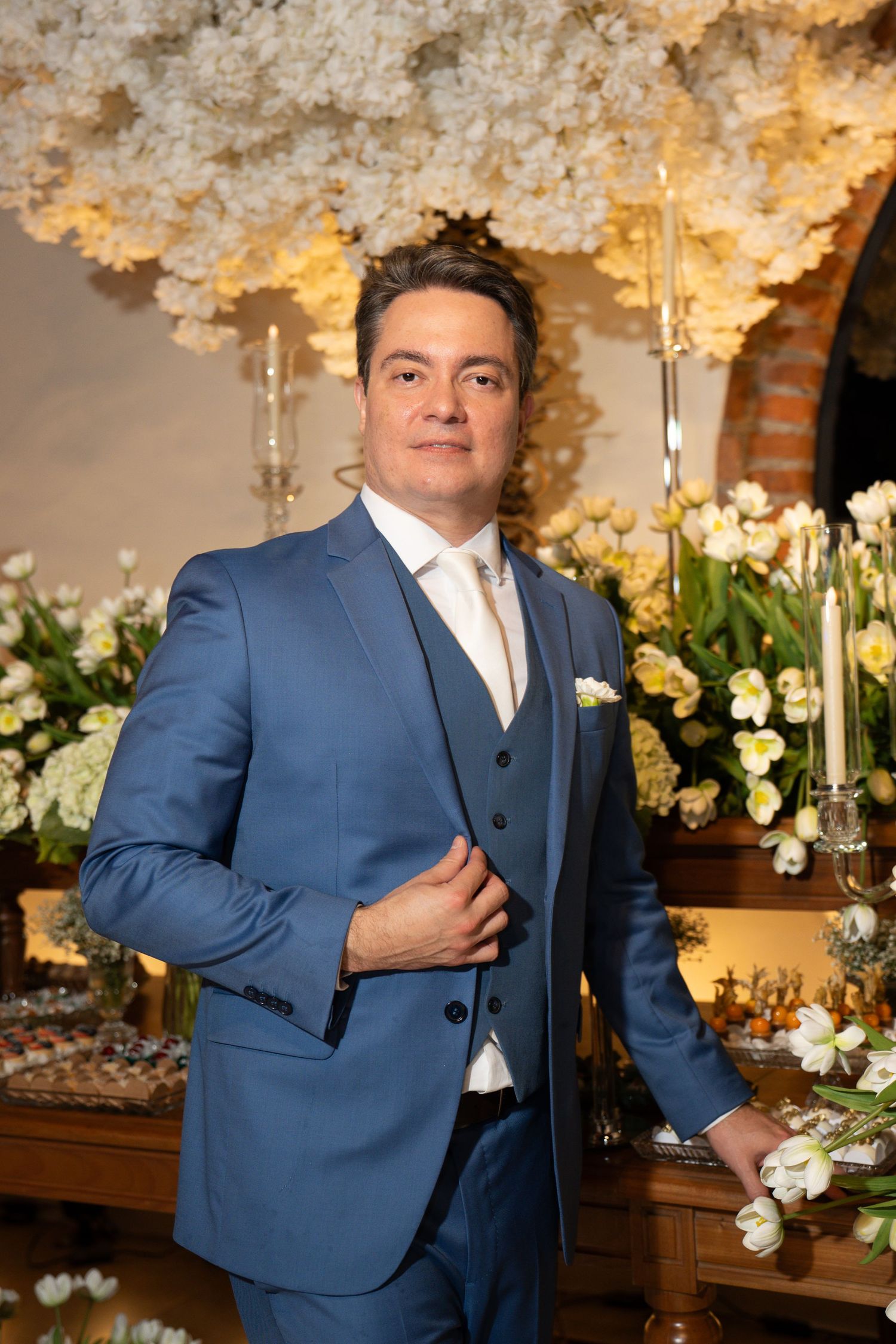Elegant man in blue three-piece suit posing at a floral wedding reception with white flower arrangements.